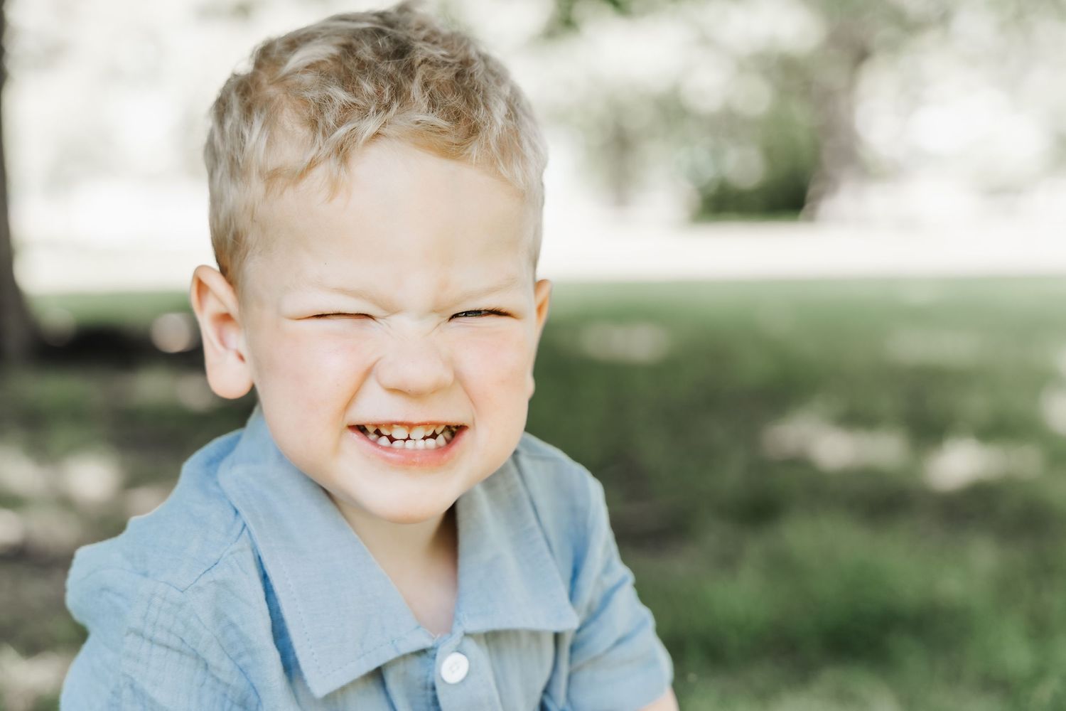 Adorable blonde toddler boy squinting and smiling outdoors in a light blue shirt.