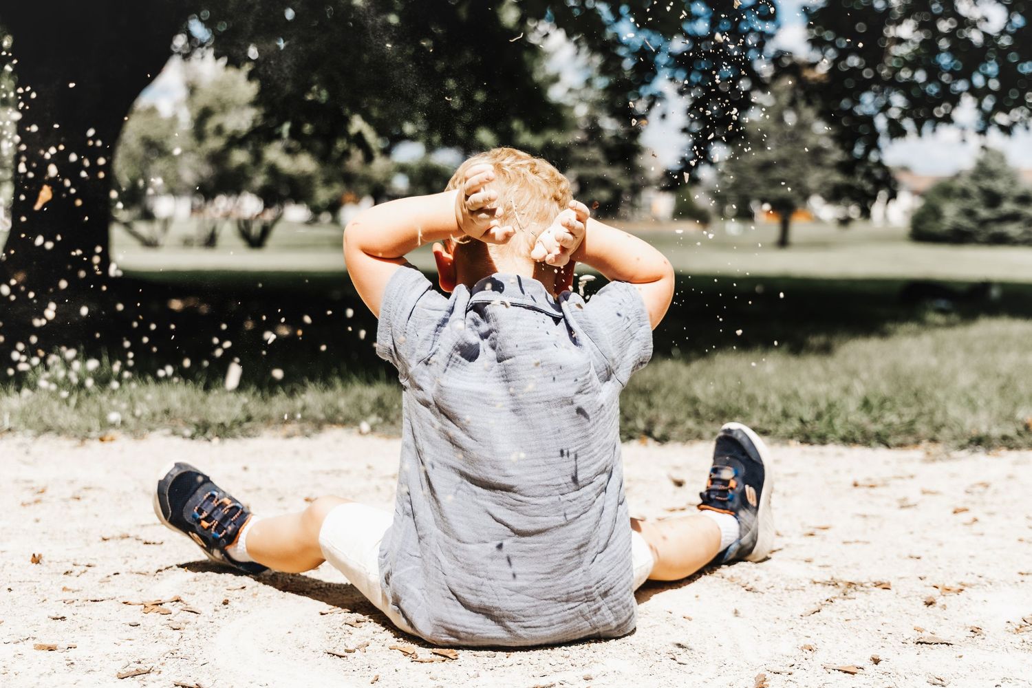 Young child sitting in sand with hands on head as confetti falls around them in a sunny park.