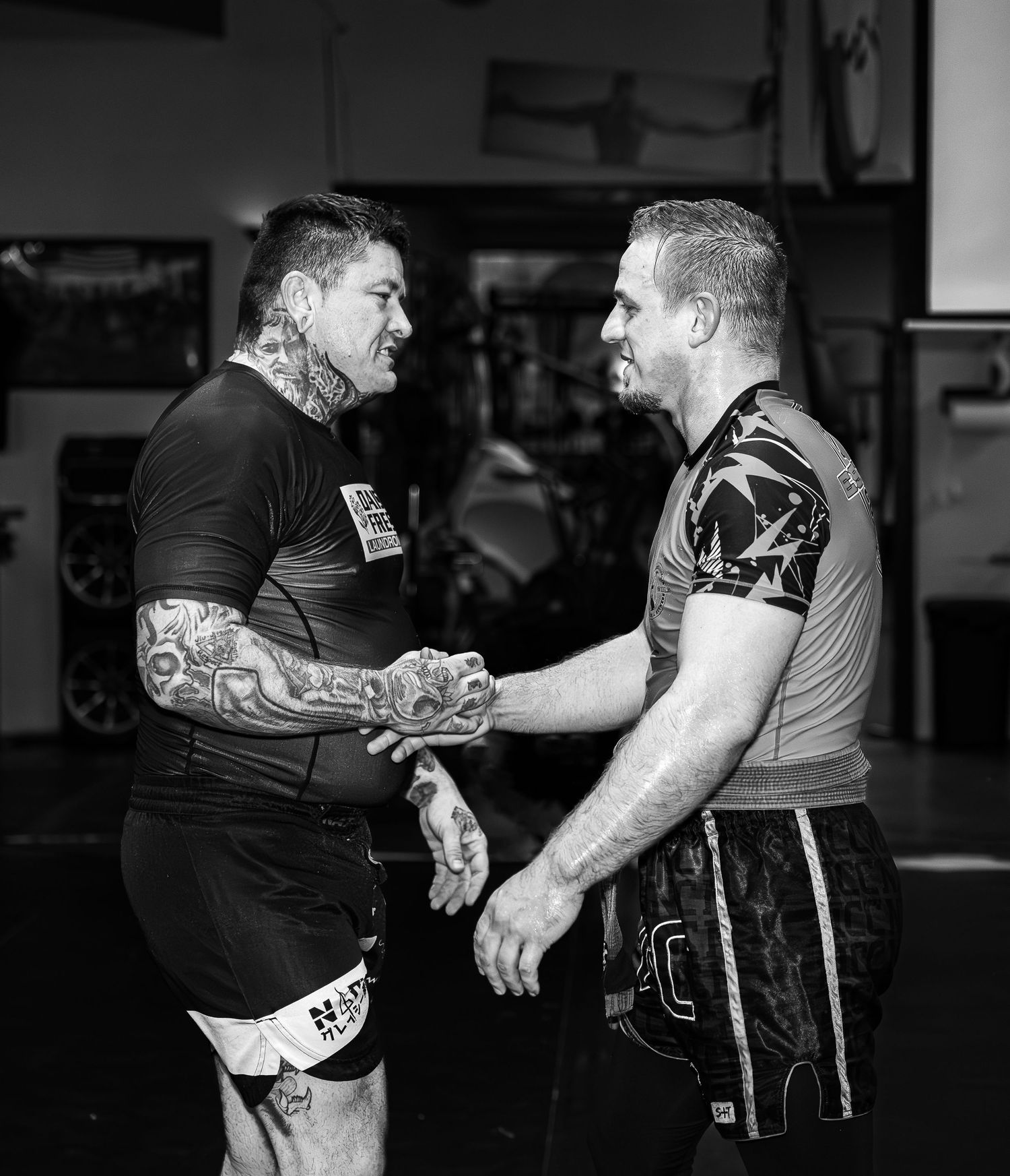 Two tattooed MMA fighters shake hands in a gym during a black and white training session.