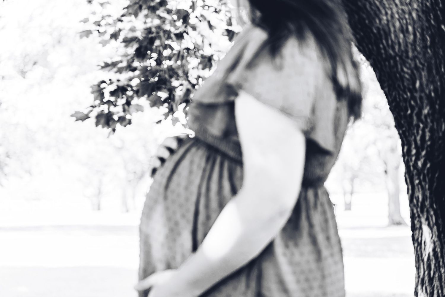Black and white profile photo of a pregnant woman in a dress standing near a tree.