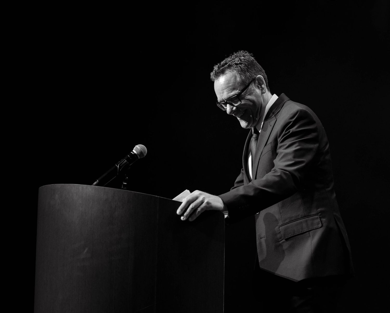 A man in a suit speaks at a podium in a dramatic black and white stage photo.