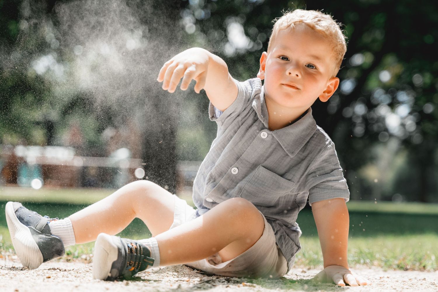 Blonde toddler boy sitting in sand at a playground, tossing sand into the air on a sunny day outdoors.