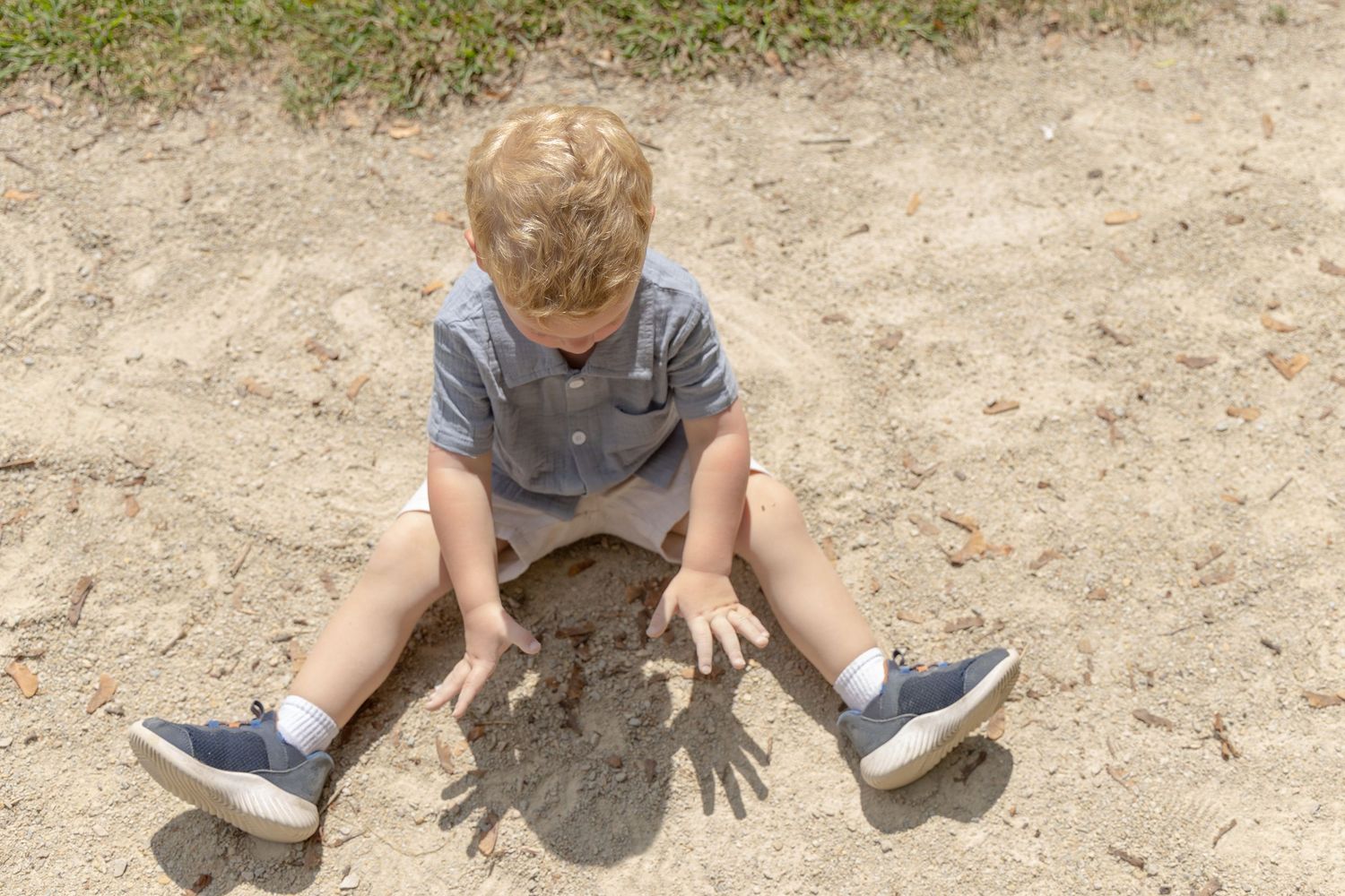 Young blonde boy sitting in sandy dirt outdoors, looking down at his hands.