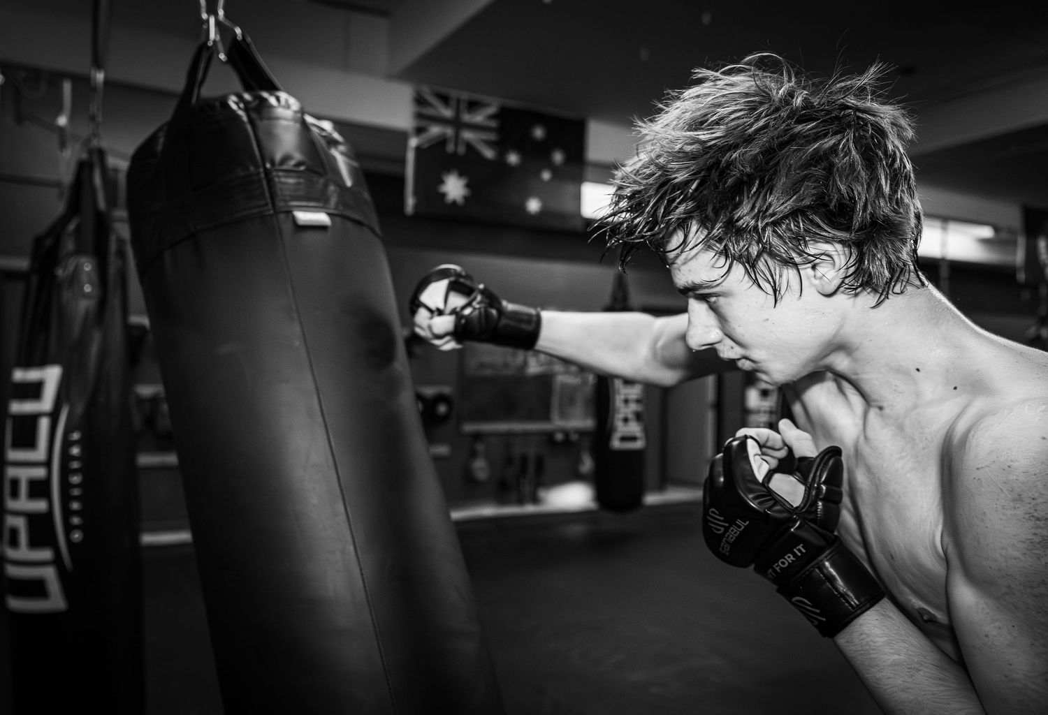 Black and white photo of a shirtless male boxer throwing punches at a heavy bag in a gym.