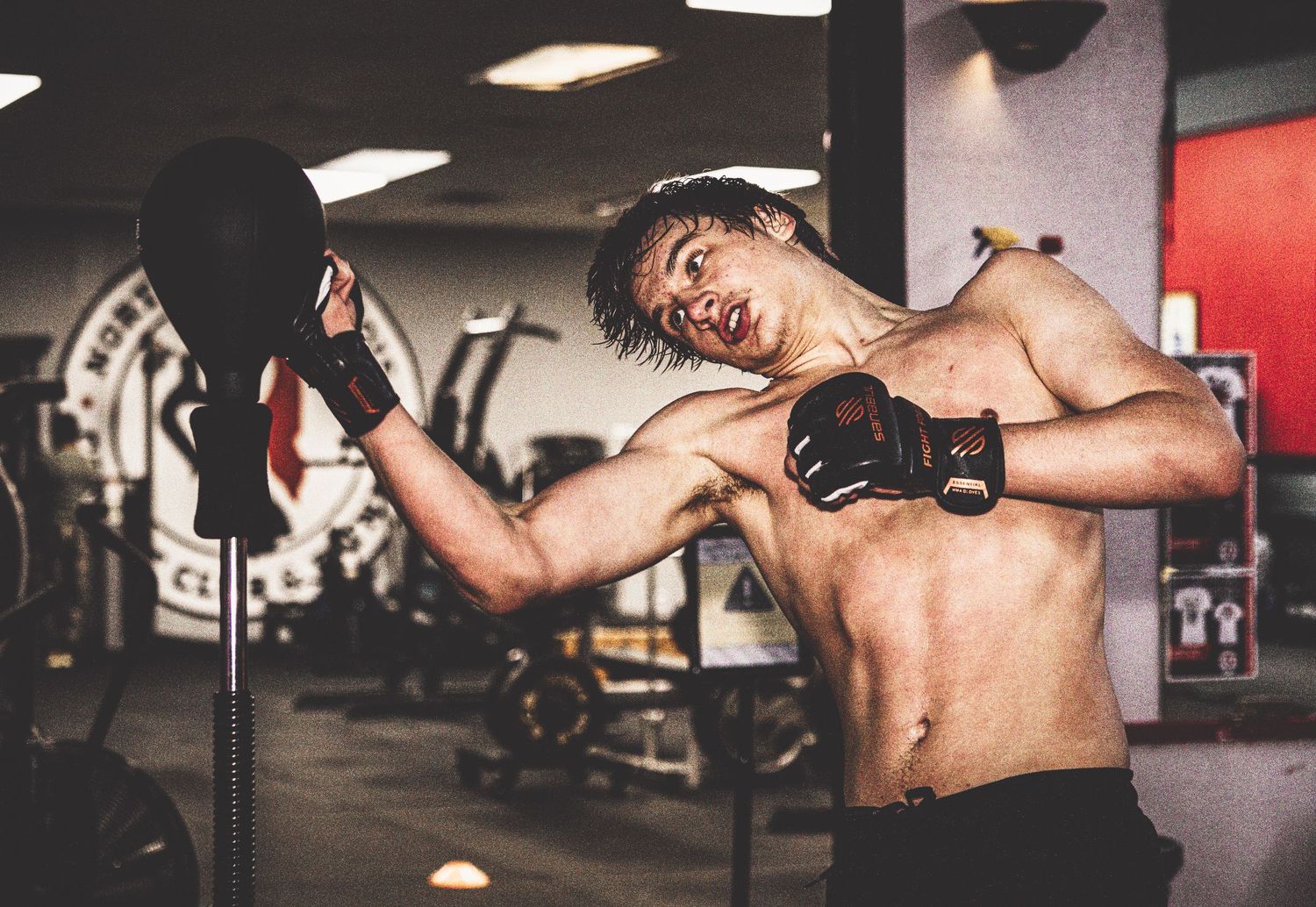 Shirtless MMA fighter training with punching pad in a gym.