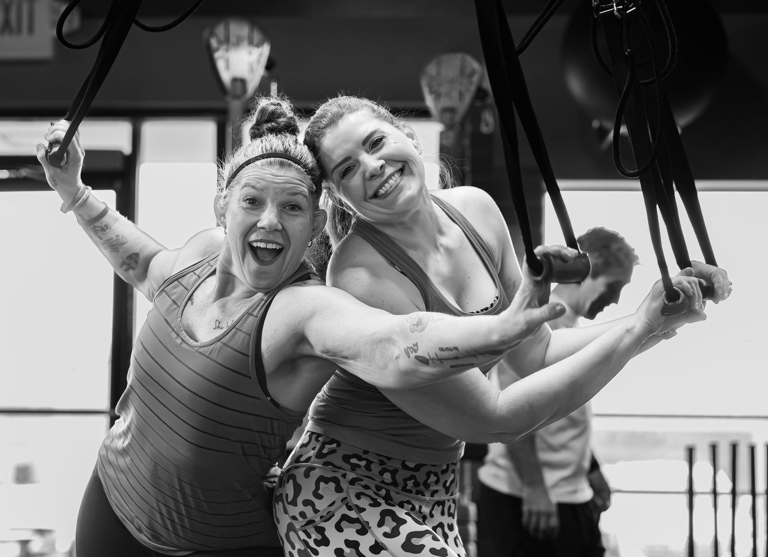 Two smiling women pose together while using suspension straps in a gym.