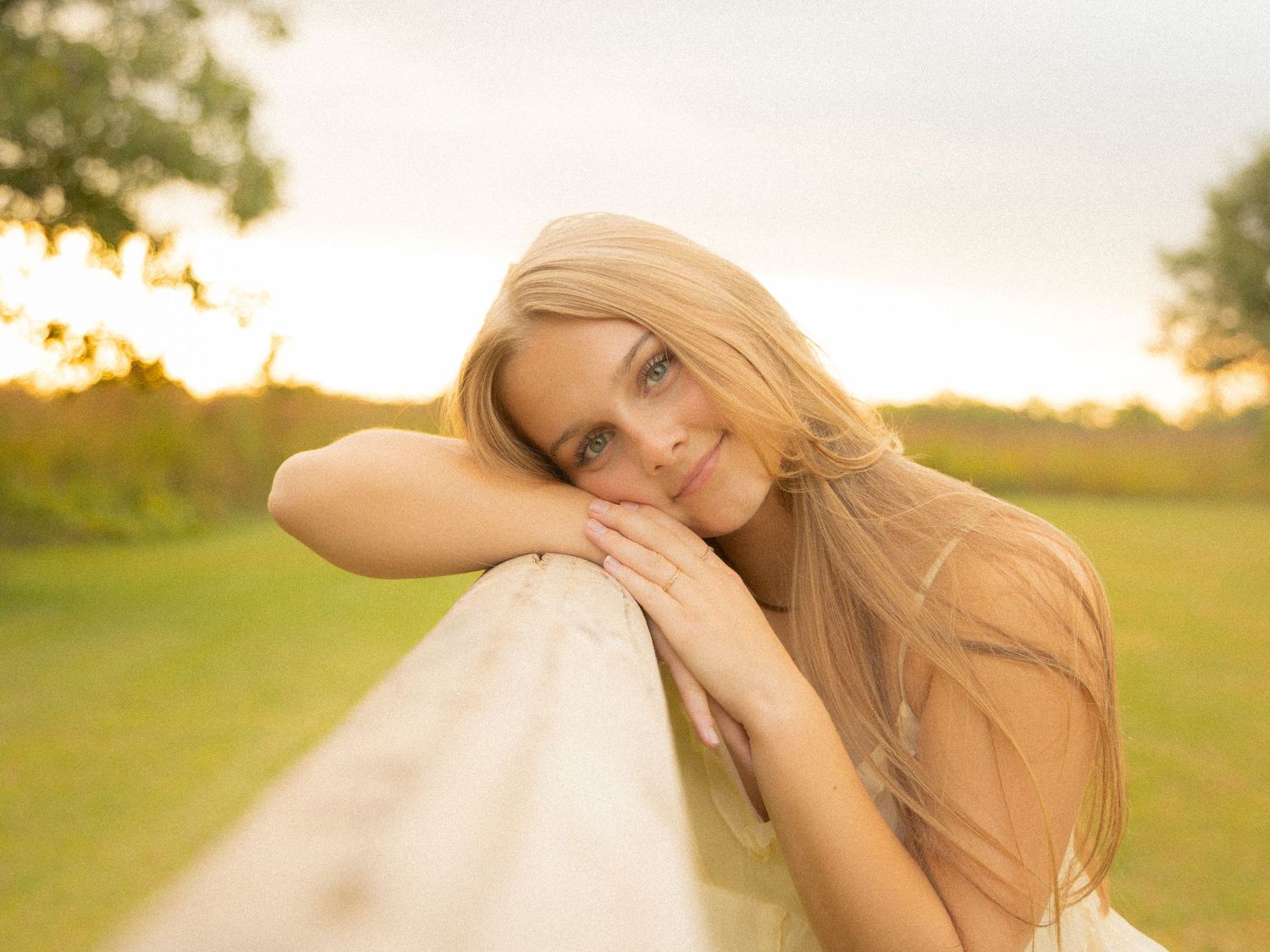 Blonde woman resting chin on white fence rail smiling at golden hour sunset.