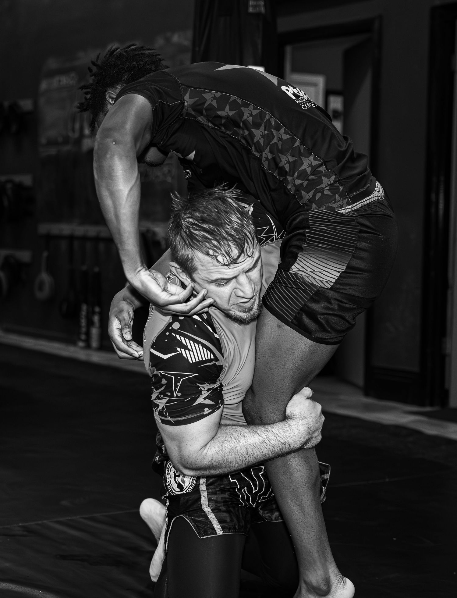 Two wrestlers grapple in black and white, one lifting the other during intense training.
