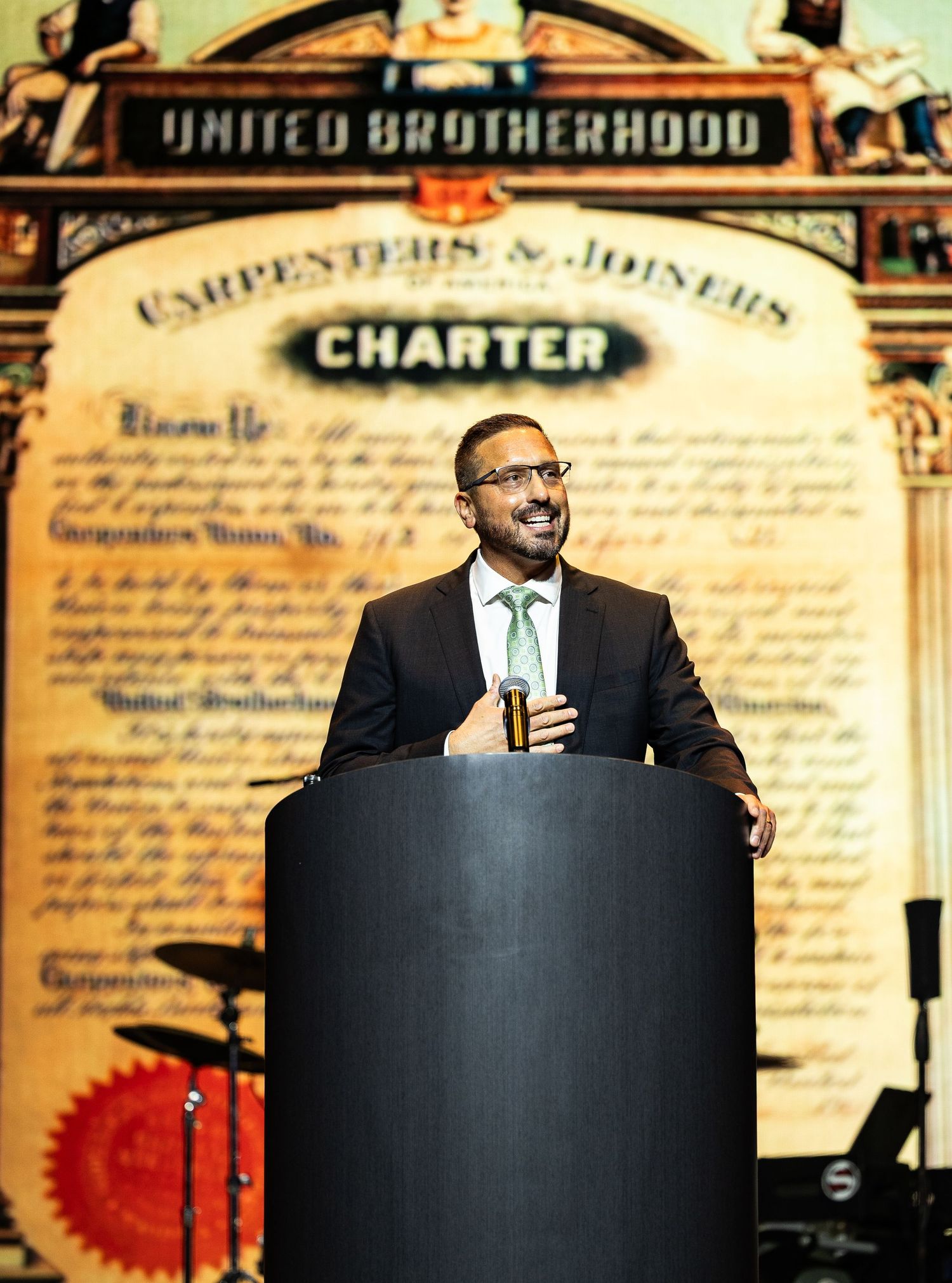 A man speaks at a podium in front of a United Brotherhood Carpenters and Joiners Charter backdrop.