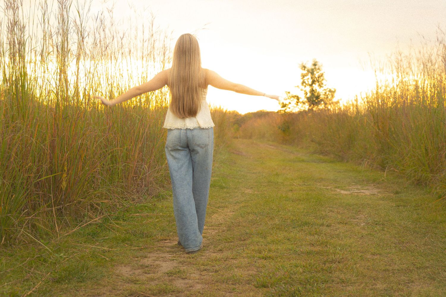 Blonde woman walking dirt path through tall grass with arms outstretched at sunset.