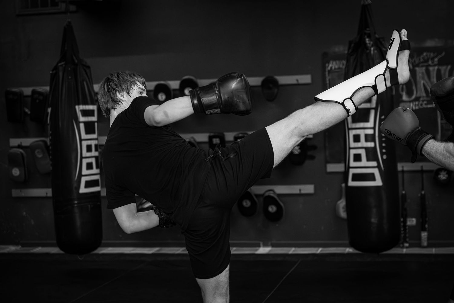 Black and white photo of a martial artist executing a high kick beside heavy bags in a gym.