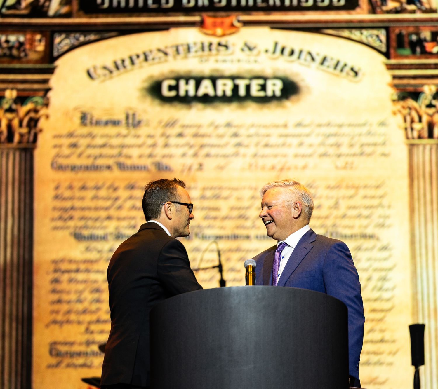 Two men shake hands at a podium in front of a United Brotherhood Carpenters and Joiners Charter backdrop.