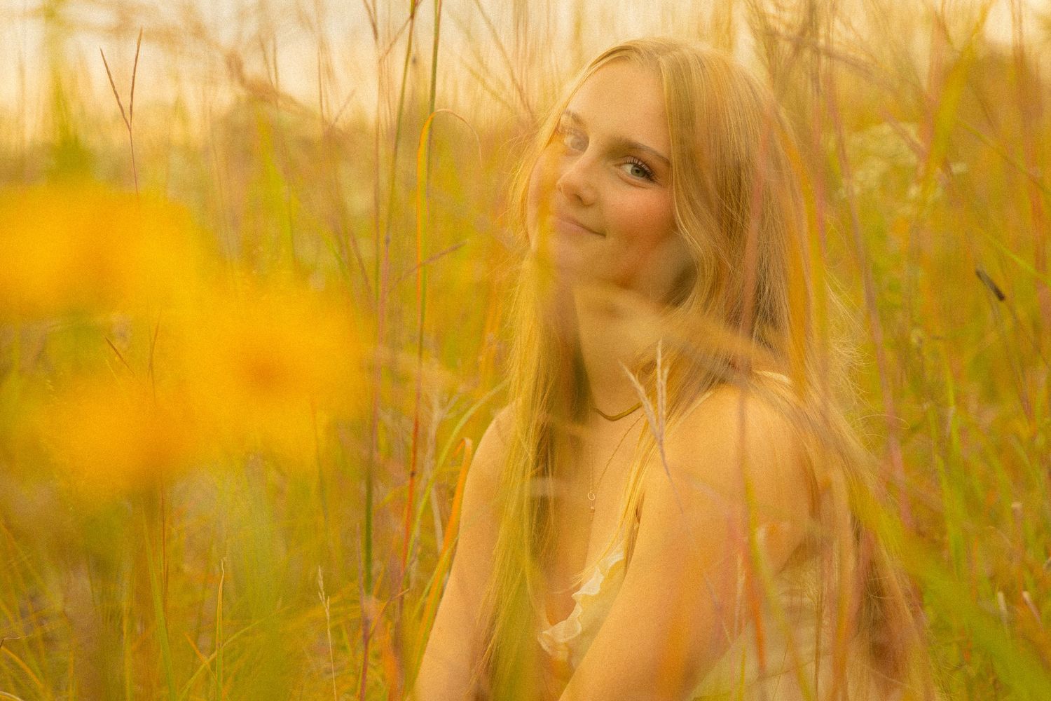 Young blonde woman smiling softly among tall golden wildflowers at sunset.