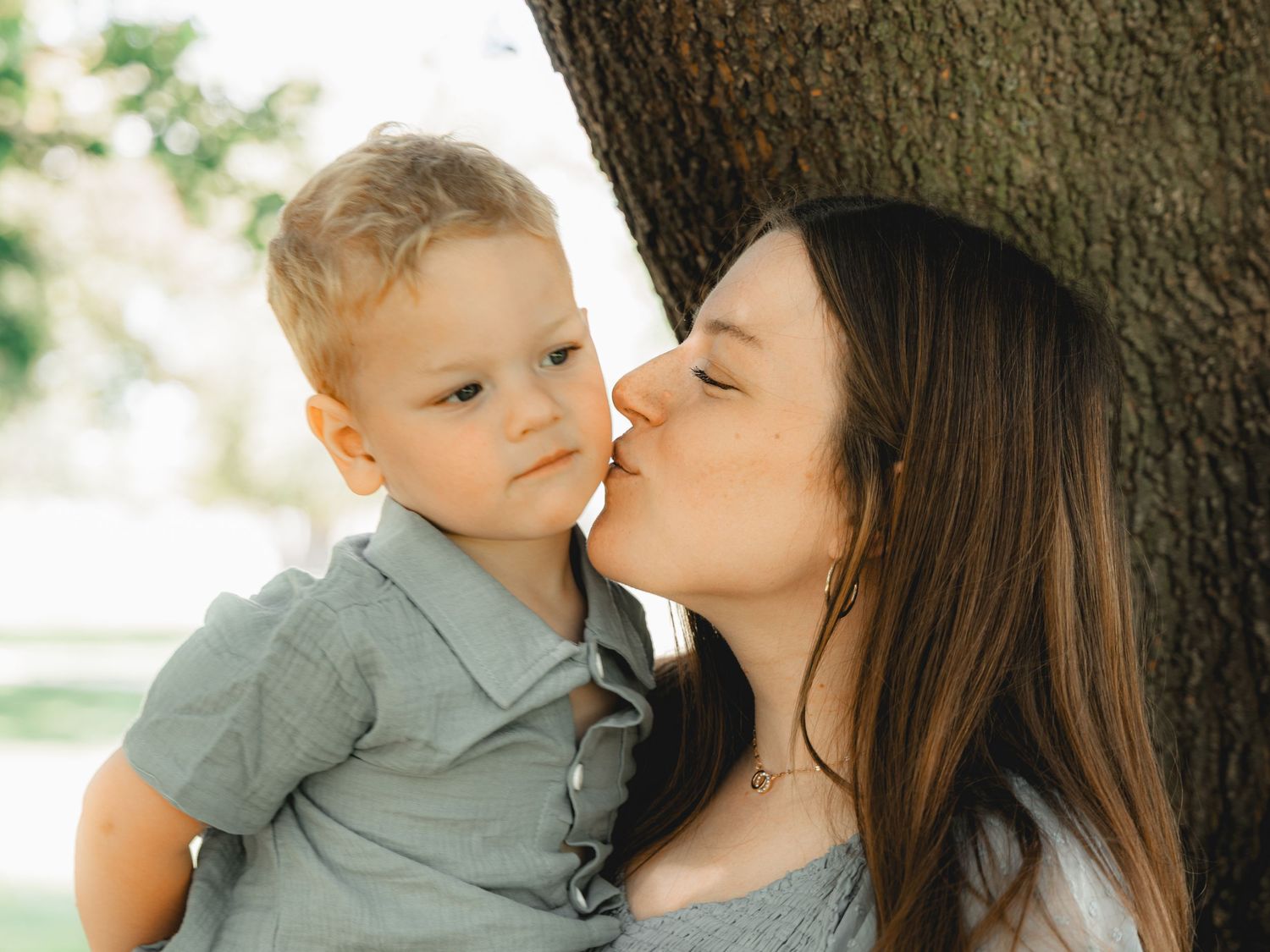 Young mother kissing her toddler son on the cheek while leaning against a tree.