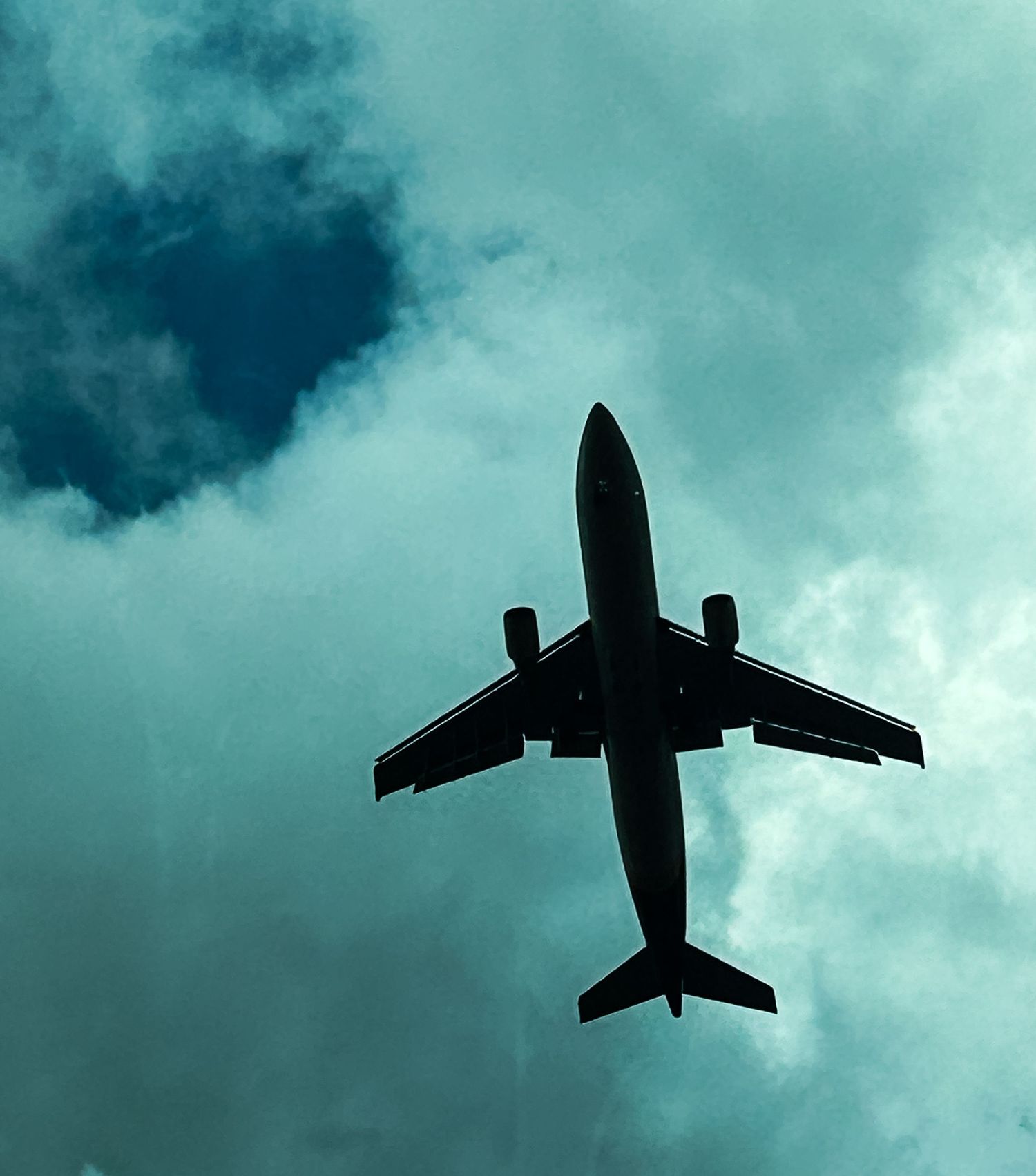 Silhouette of a commercial airplane flying overhead against a dramatic teal cloudy sky, viewed from below.