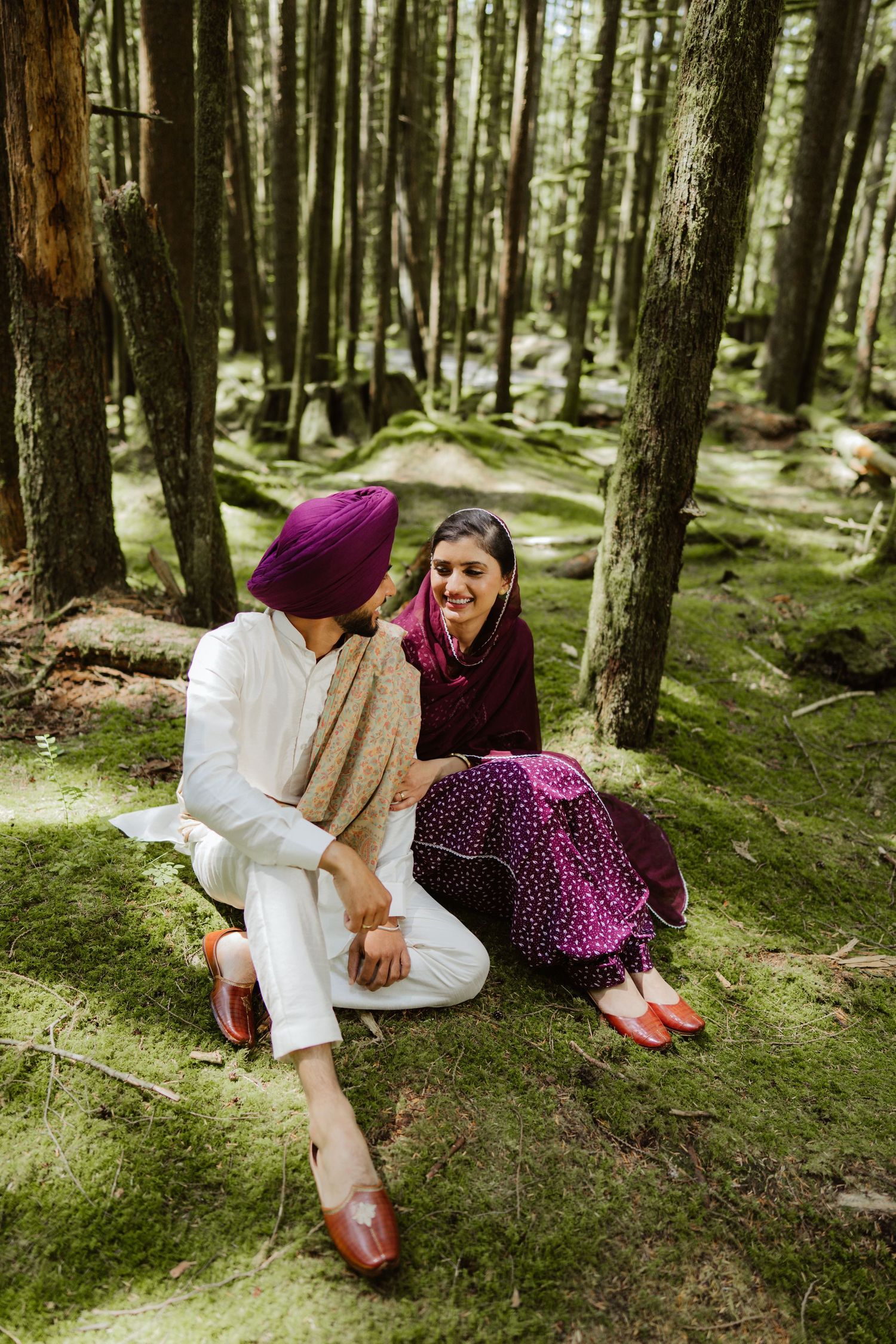 Sikh couple laughing and sitting on mossy ground in a lush green forest setting.