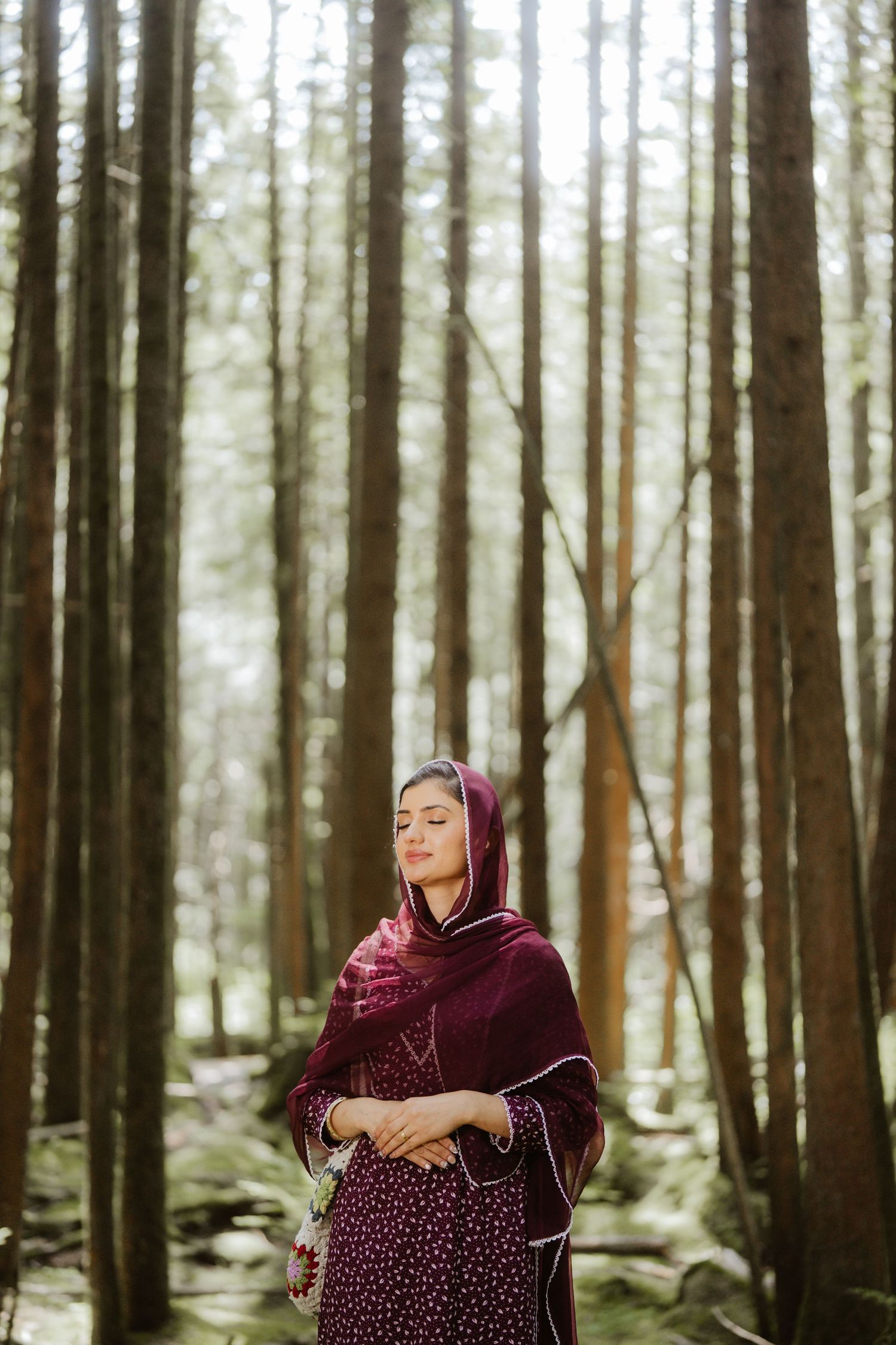 A woman in a burgundy traditional South Asian outfit stands serenely in a sunlit pine forest.