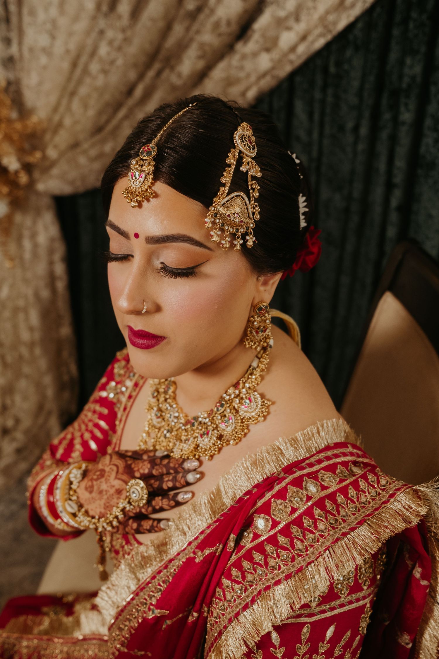 Indian bride in red saree with gold jewelry, maang tikka, and mehndi, looking down gracefully.