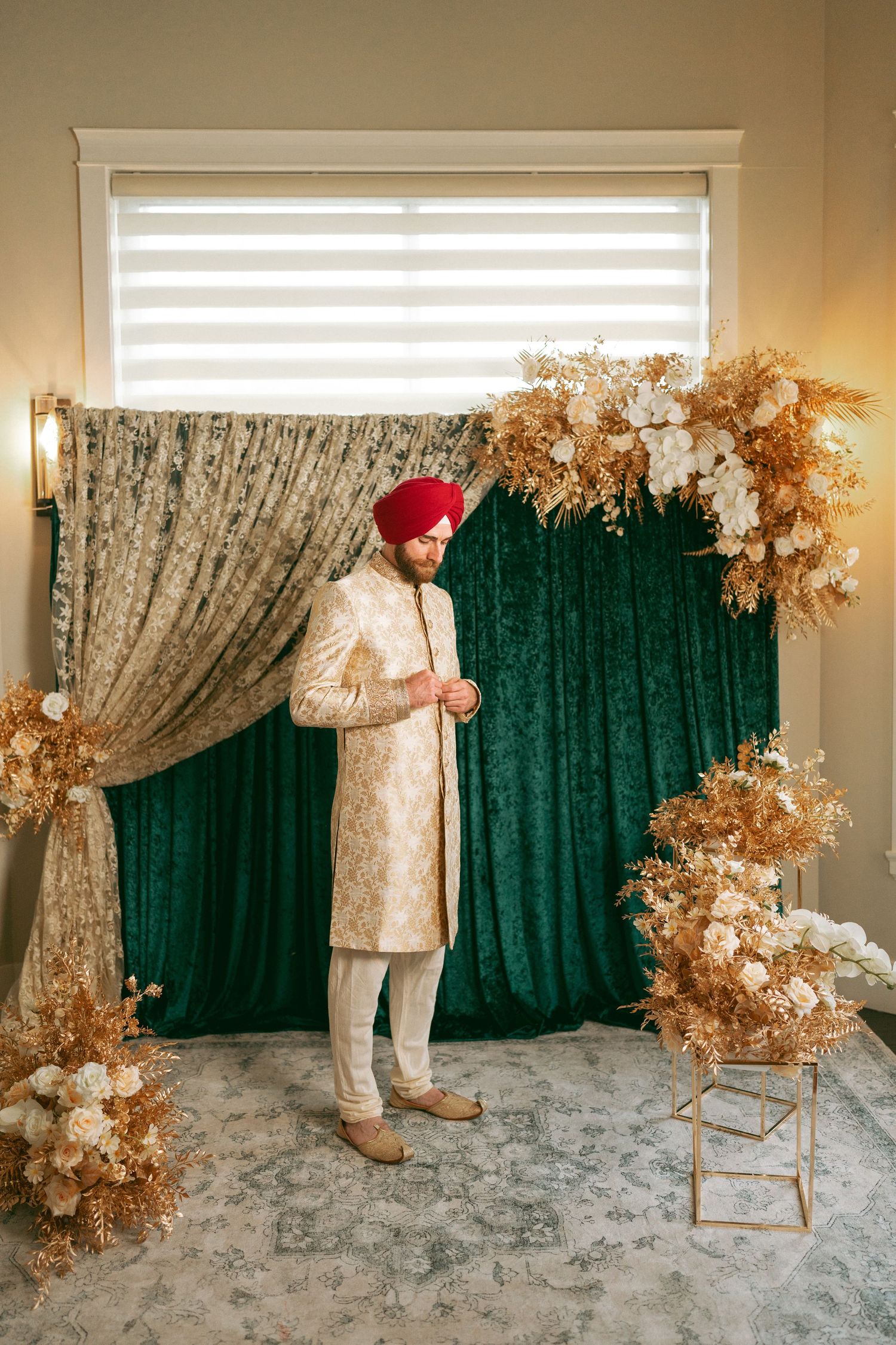 Sikh groom in red turban and ivory sherwani stands before green velvet backdrop with golden floral arrangements.