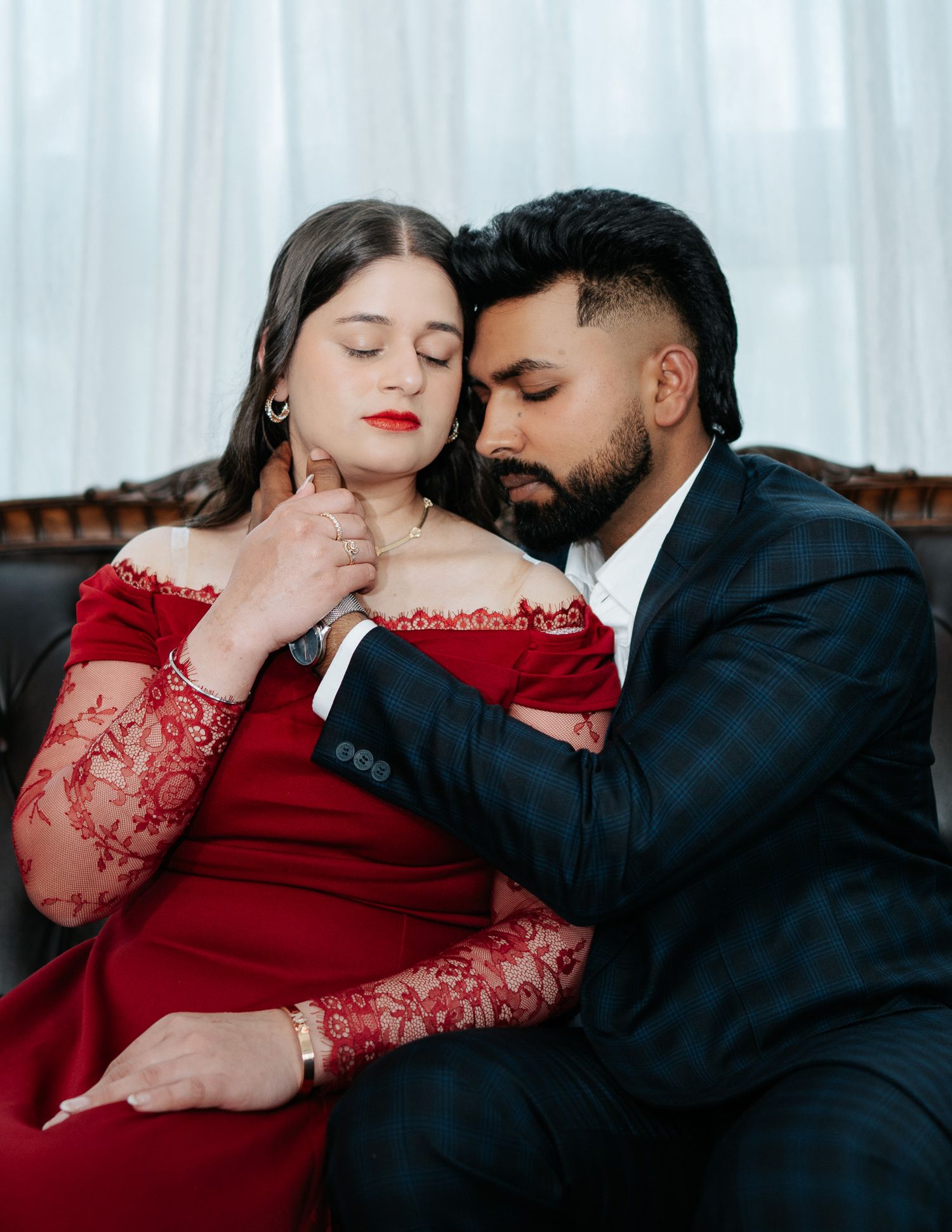 Romantic couple posing together, woman in red lace dress with man in plaid suit on leather sofa.