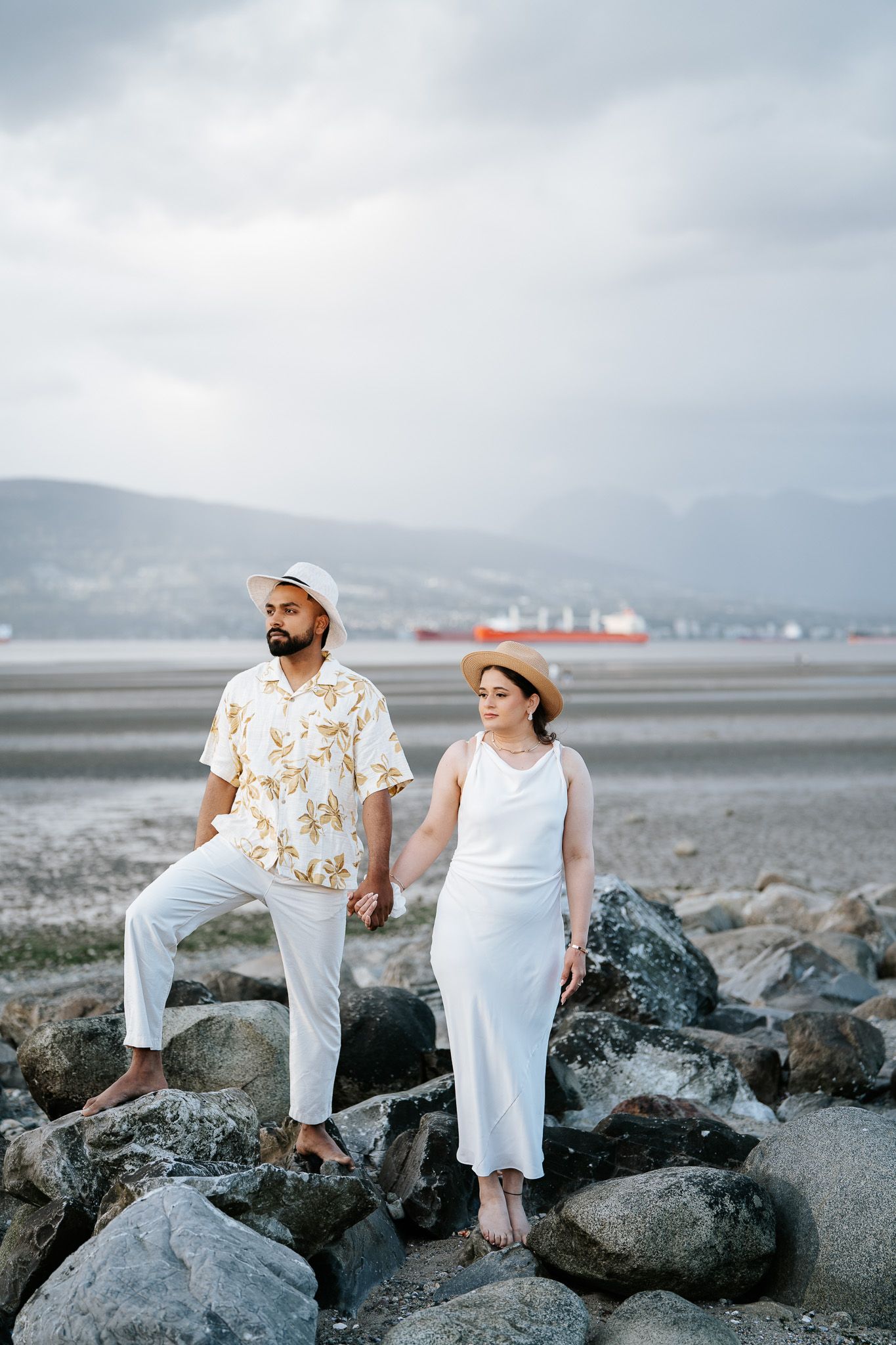 Couple holding hands on rocky beach, man in floral shirt and white pants, woman in white dress and straw hat, cloudy sky.