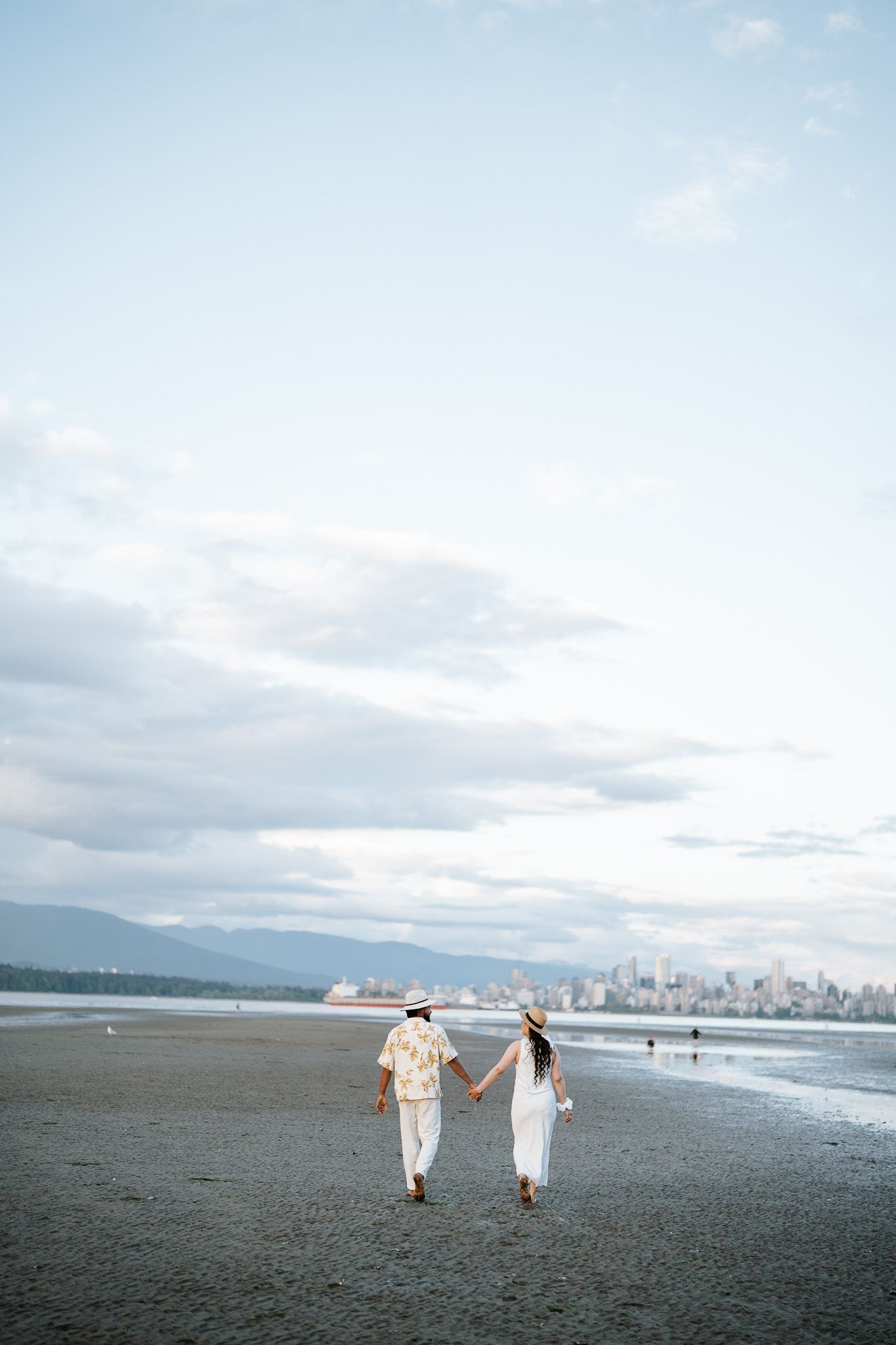 Couple holding hands walking on a sandy beach with a city skyline and mountains in the background.