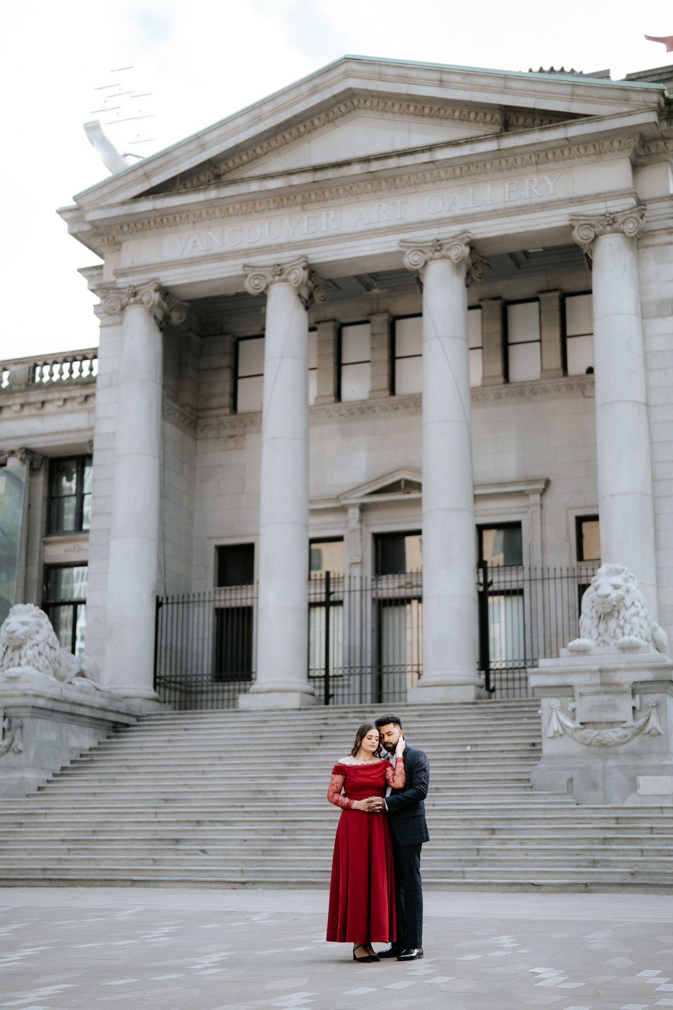 Couple embracing in front of grand neoclassical building with columns and lion sculptures on steps.