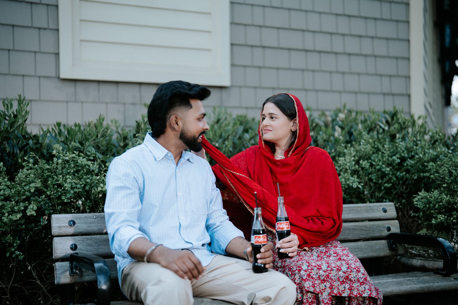 Couple sitting on outdoor bench sharing drinks, woman in red hijab and floral outfit smiling at man.