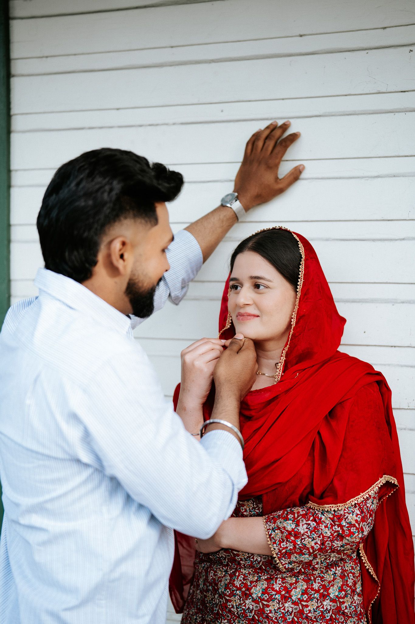 Bearded man in white shirt tenderly interacts with woman in red traditional attire against white wall.