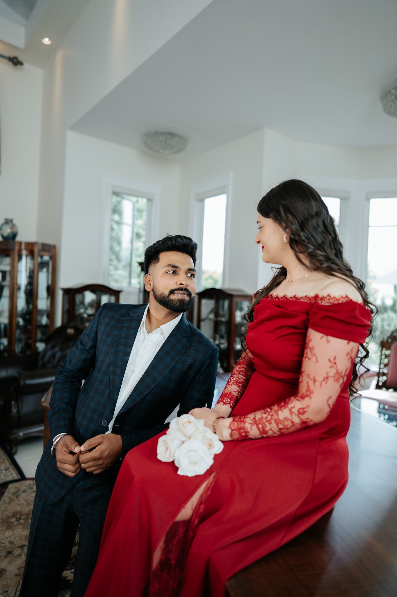 A couple in formal attire share a romantic moment, woman in red lace gown holding white roses indoors.