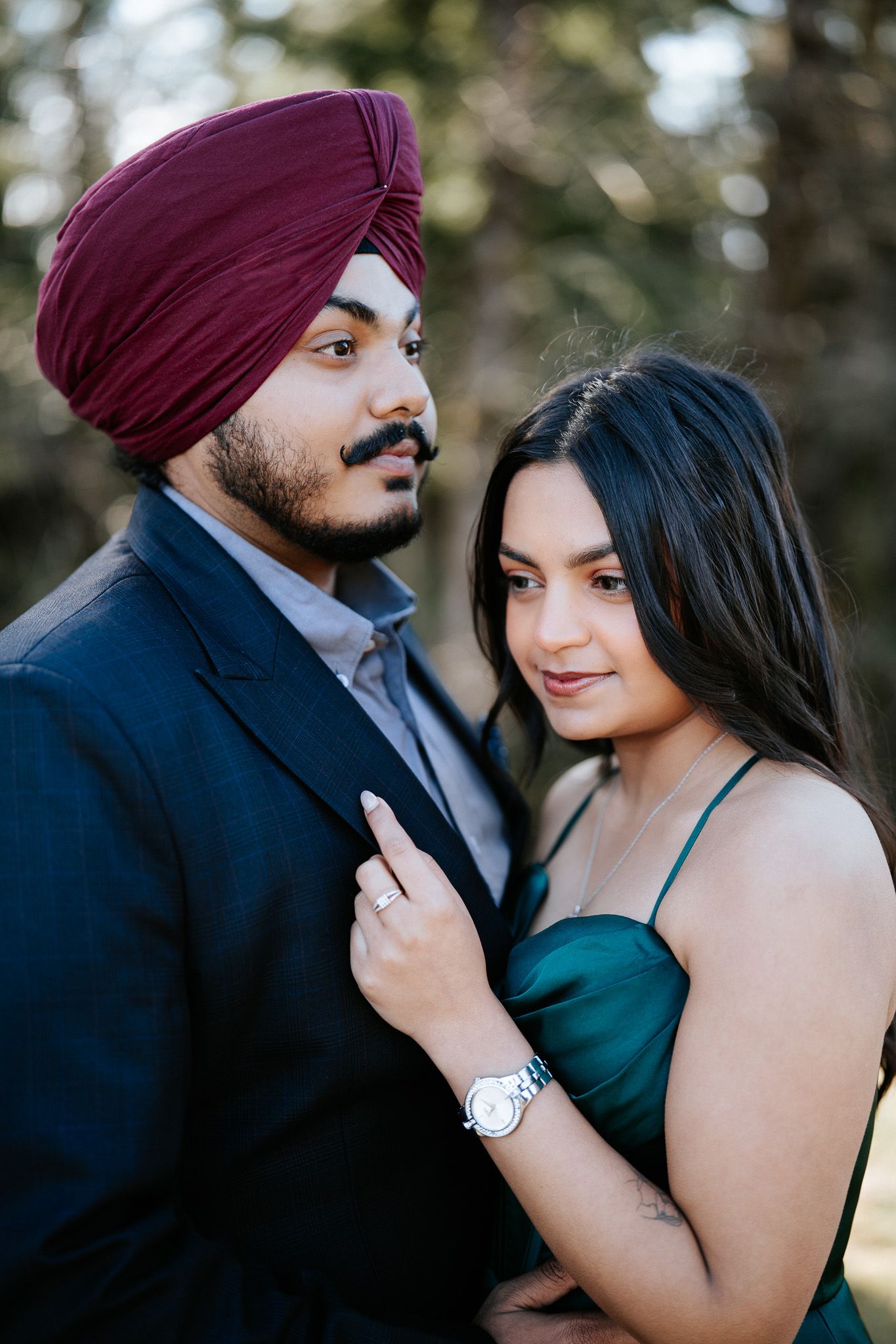 A couple poses outdoors, with one wearing a burgundy turban and navy suit while the other wears an emerald dress.
