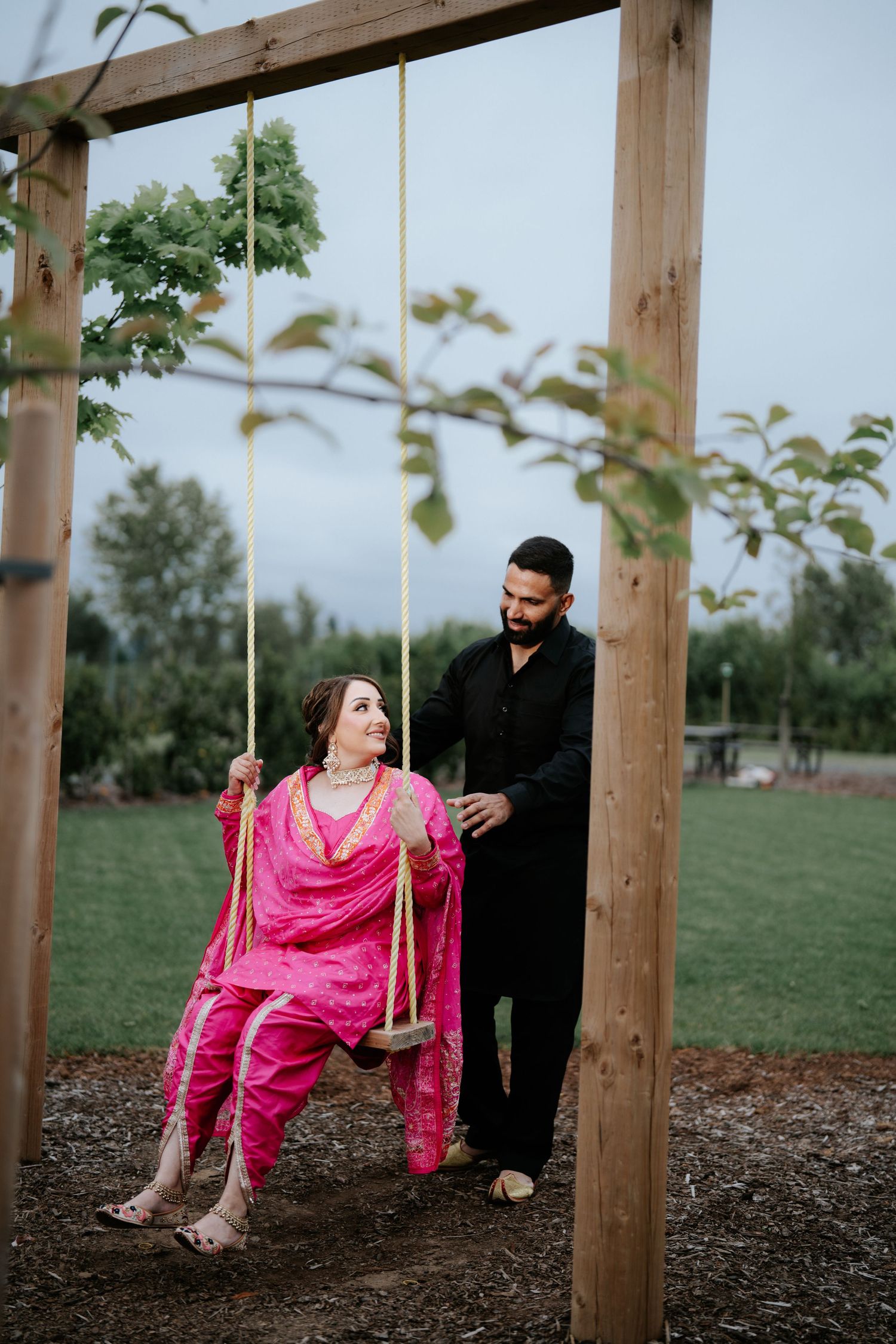 A woman in pink salwar kameez sits on a wooden swing while a man in black stands behind her outdoors.