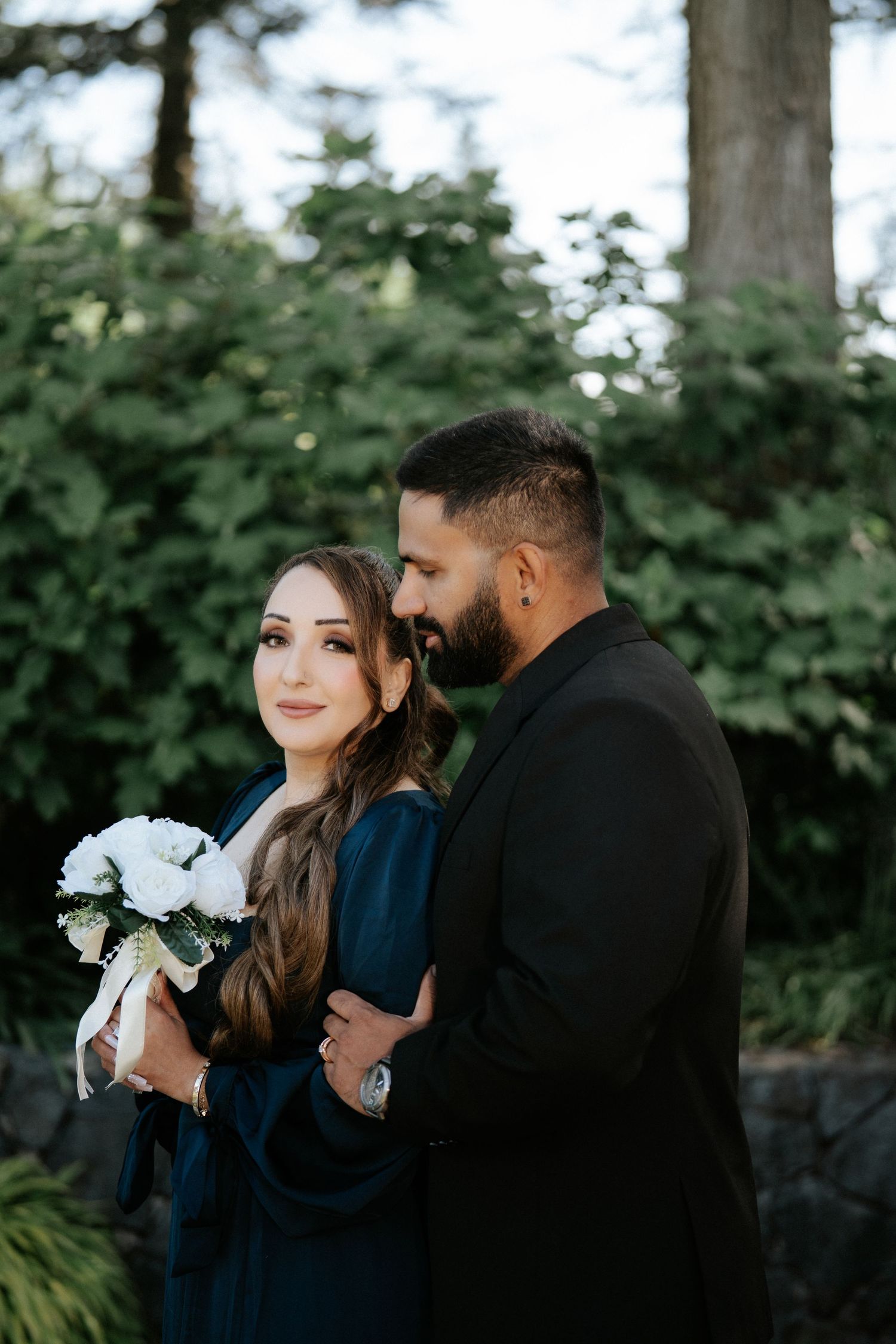 Couple in elegant attire embracing outdoors, woman in teal holding white bouquet, man in black suit, lush greenery background.