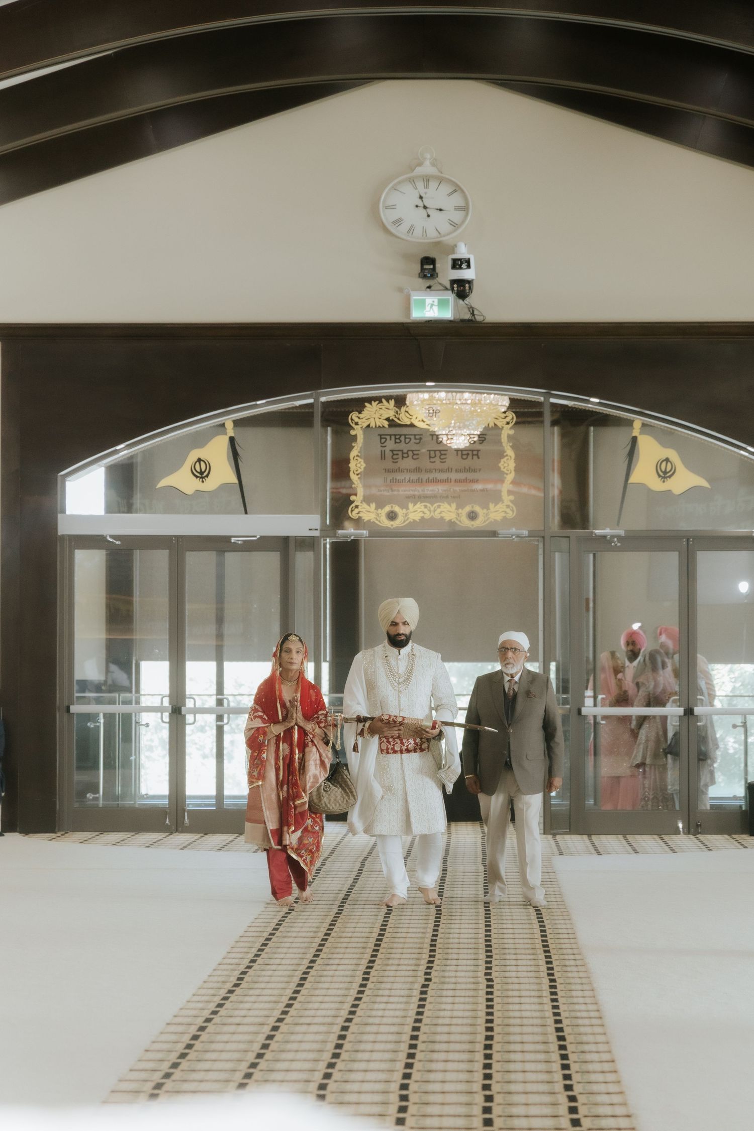 Sikh wedding couple in traditional attire walking out of a Gurdwara entrance with family members.
