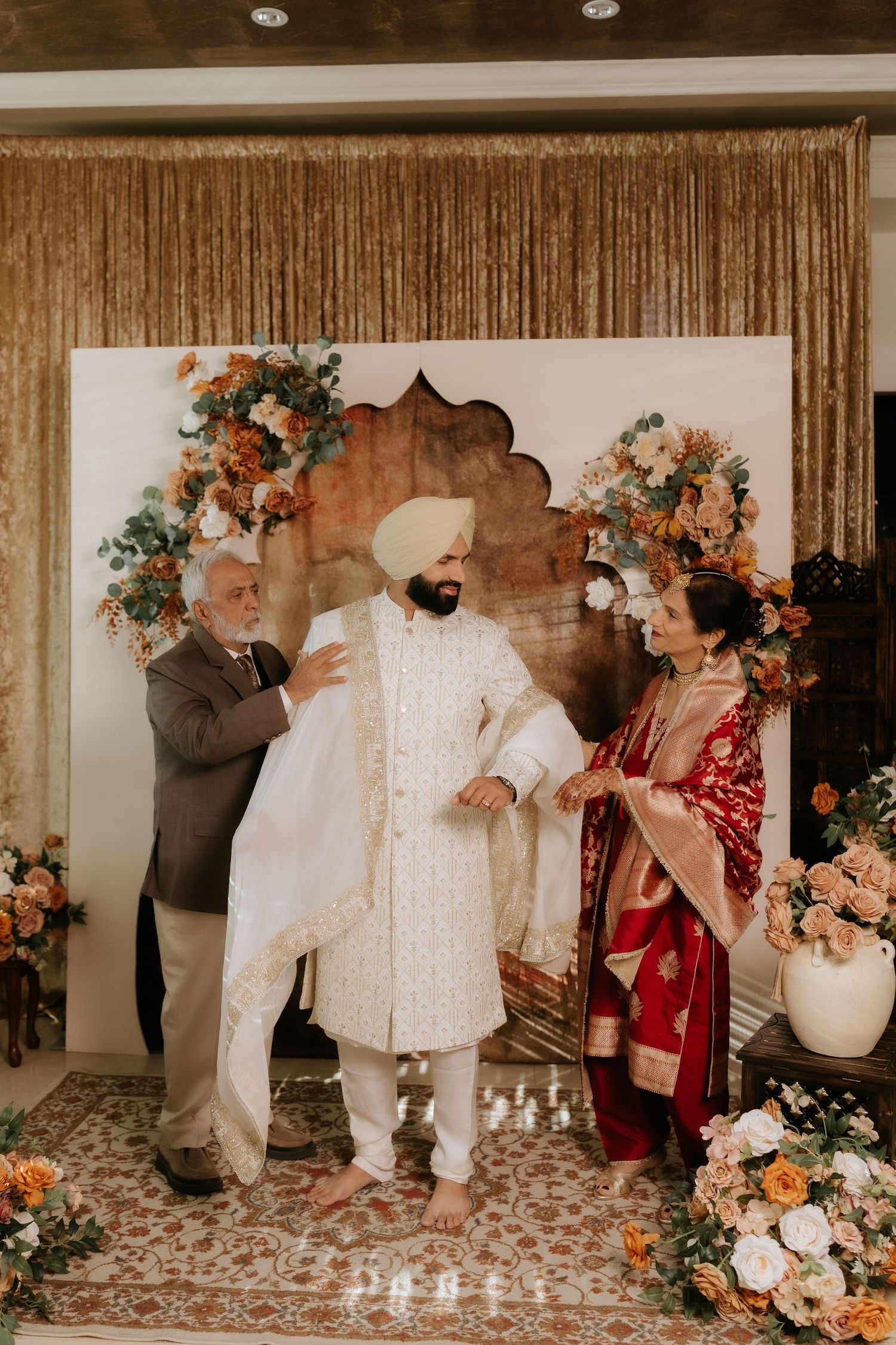 Sikh groom in white sherwani and turban with parents during traditional wedding ceremony with floral decor.