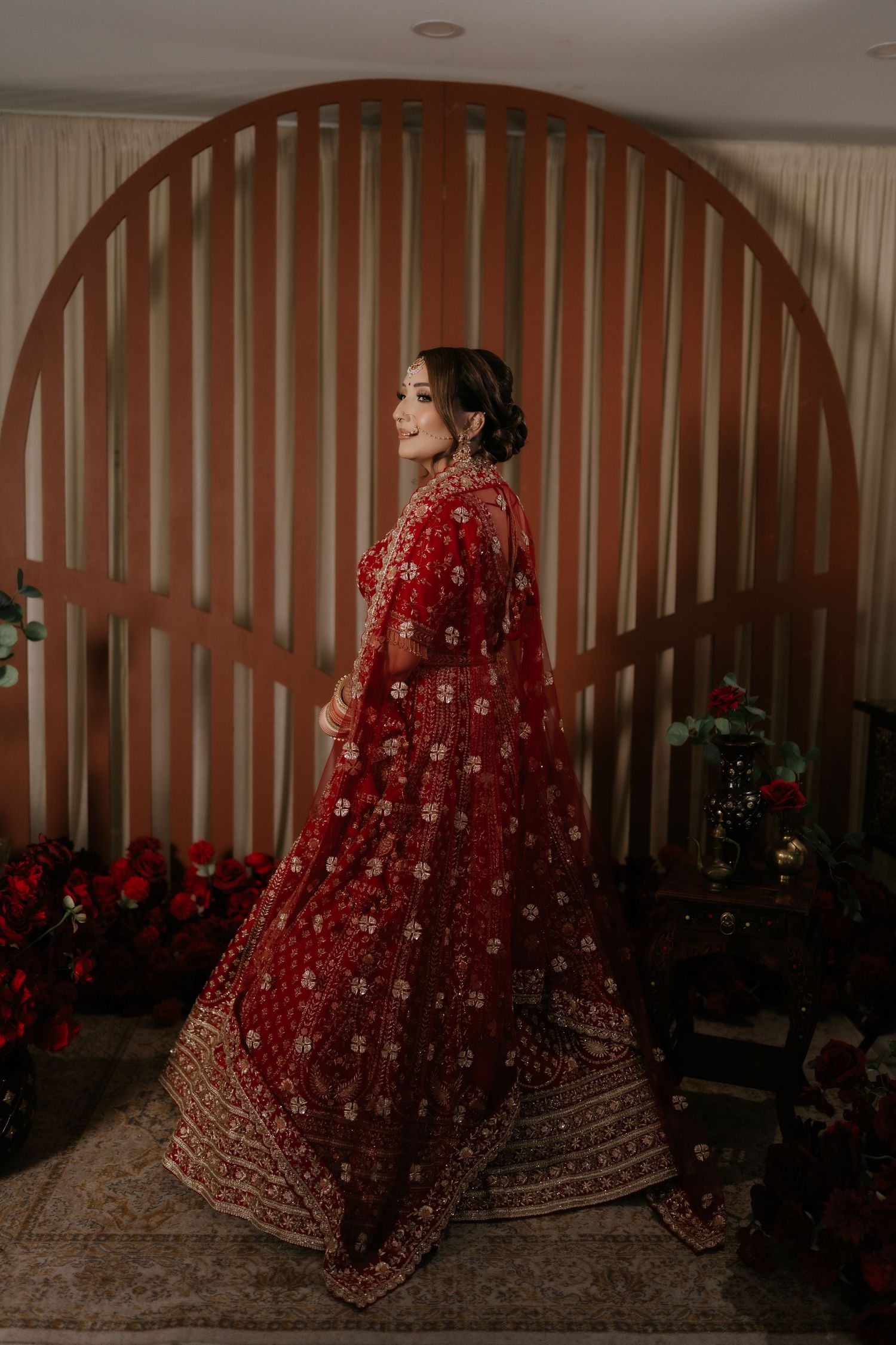 Bride in red embroidered lehenga with dupatta poses elegantly in front of a circular wooden arch backdrop.