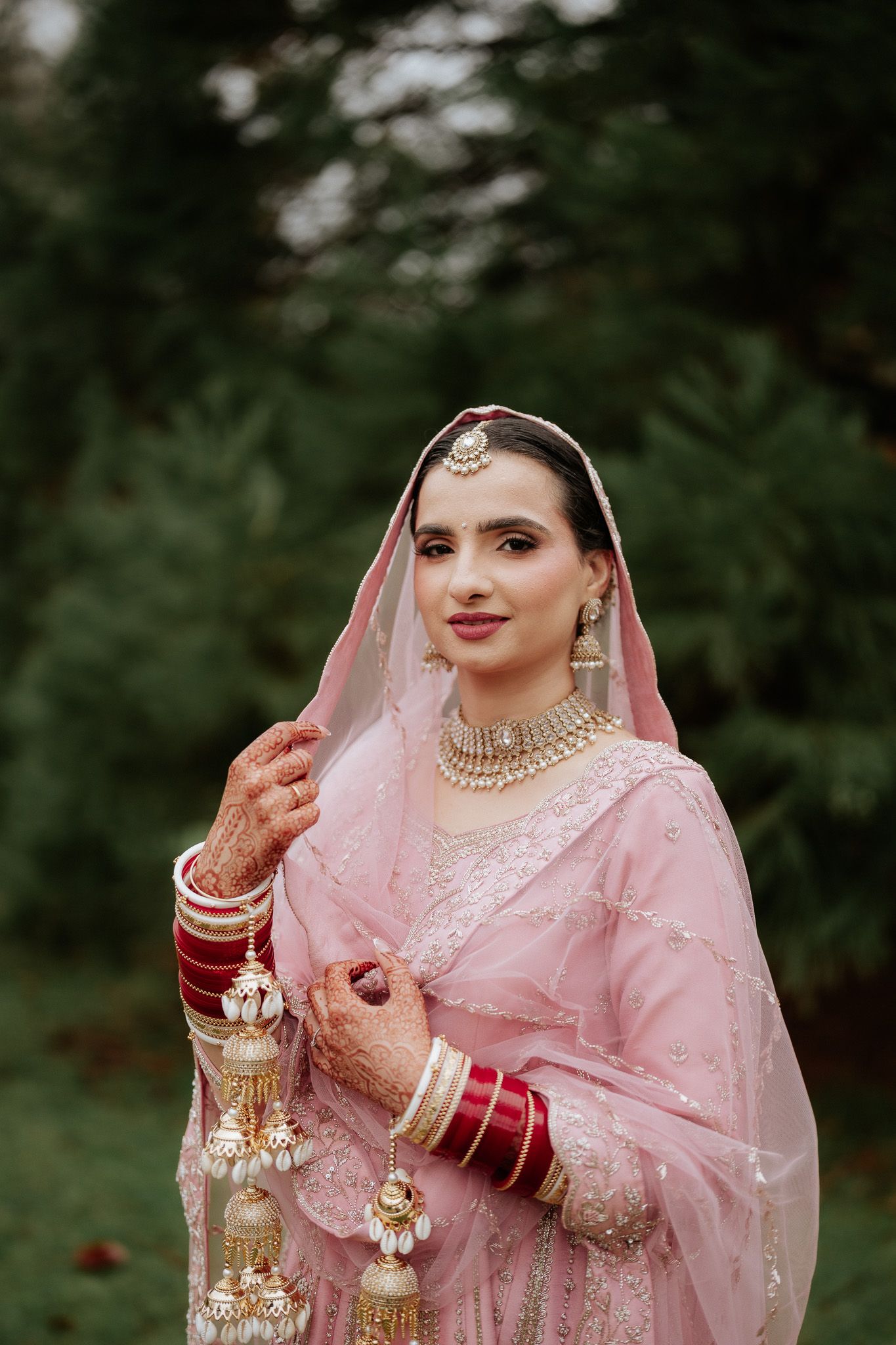 Bride in pink traditional Indian attire with gold jewelry and henna decorated hands against green foliage.