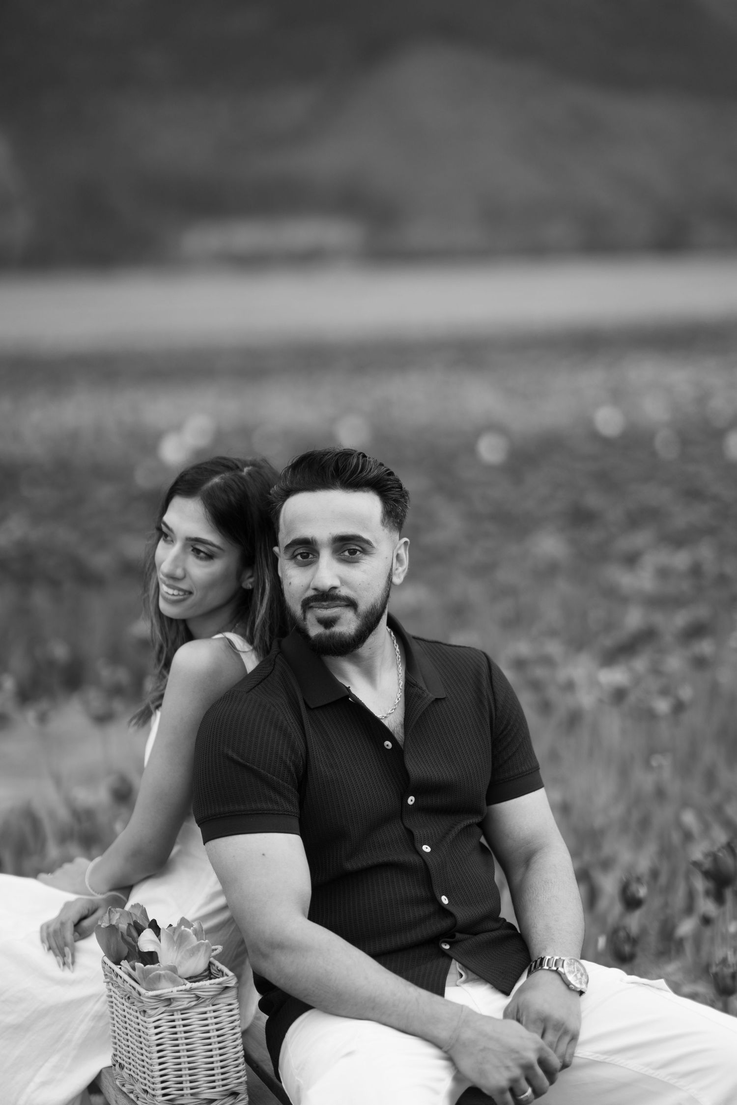A couple relaxes on a white blanket with a picnic basket in a grassy outdoor setting, captured in black and white.