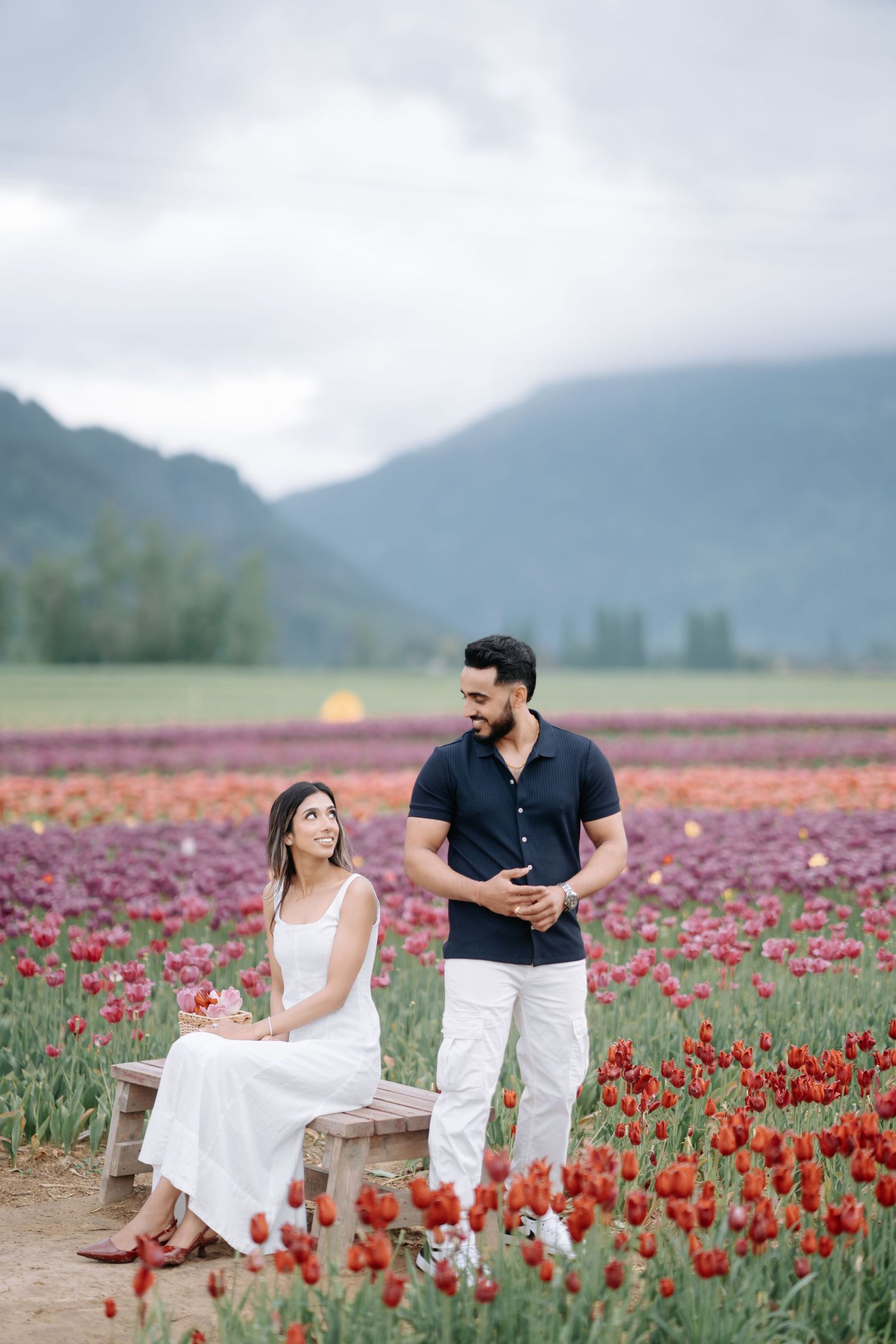 A couple stands together in a vibrant red tulip field with mountains and cloudy skies in the background.