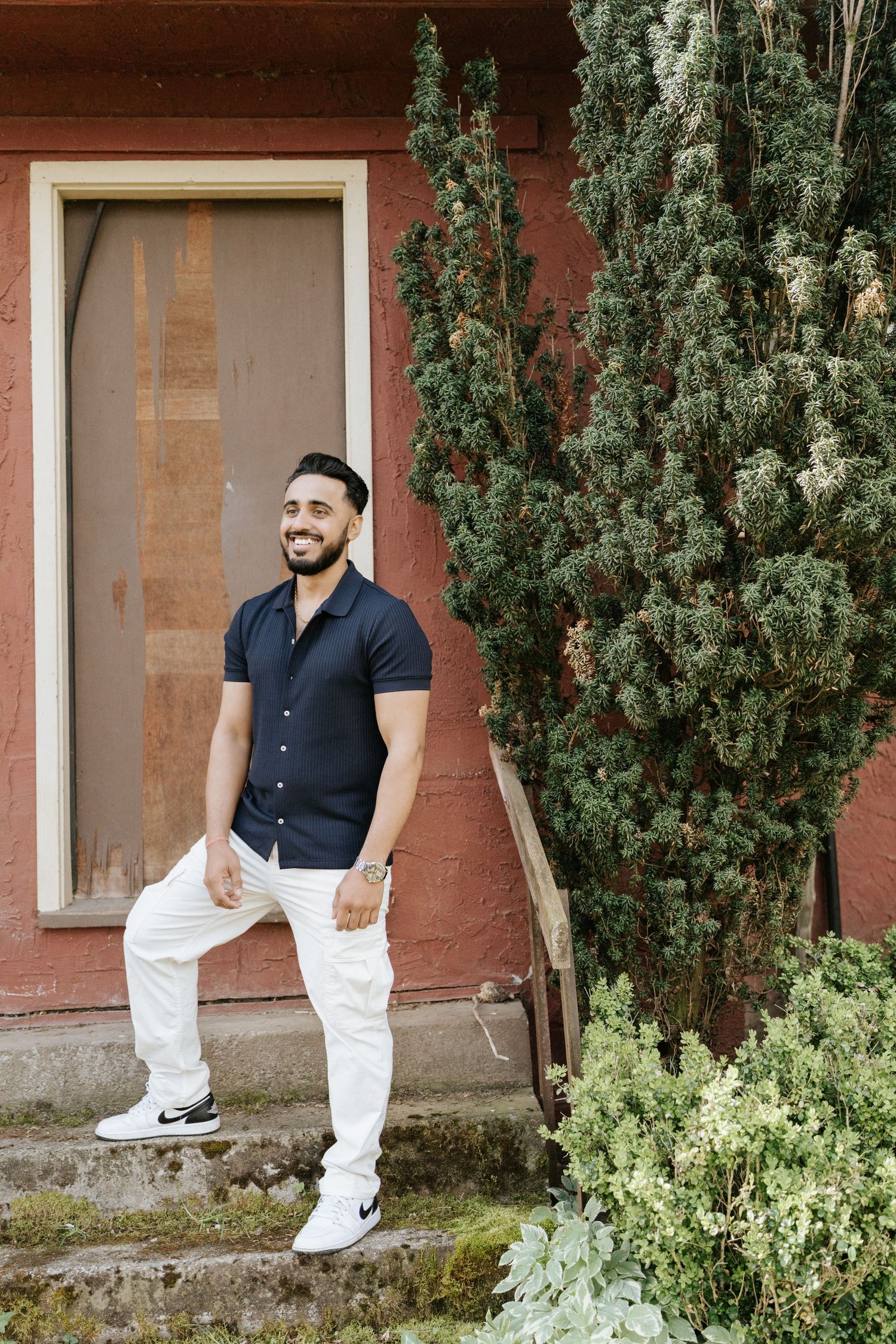 Person in navy shirt and white pants poses casually against a red building wall with green foliage.