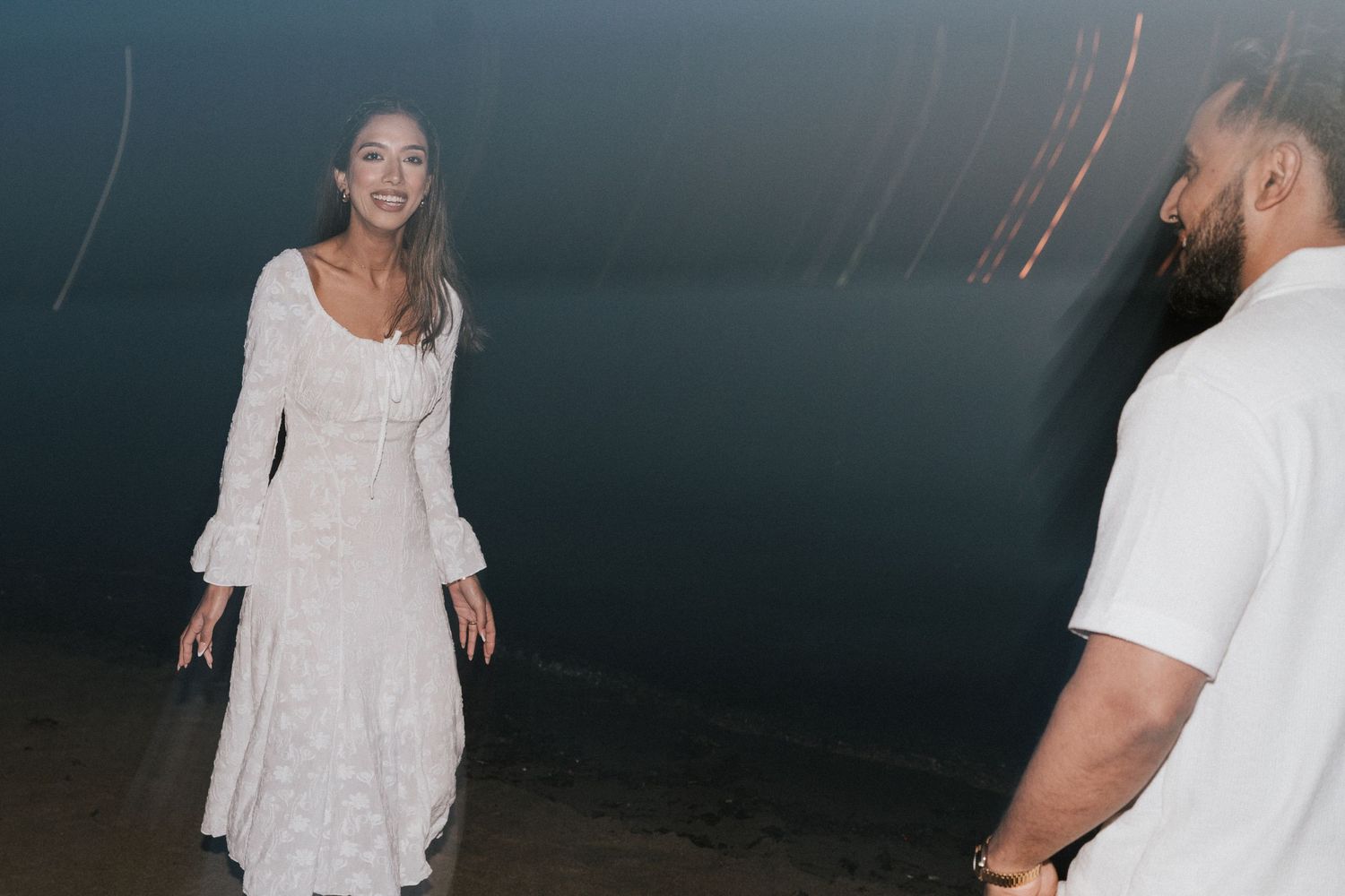 A person in a white shirt and light-colored pants walks along a dark wall at night.
