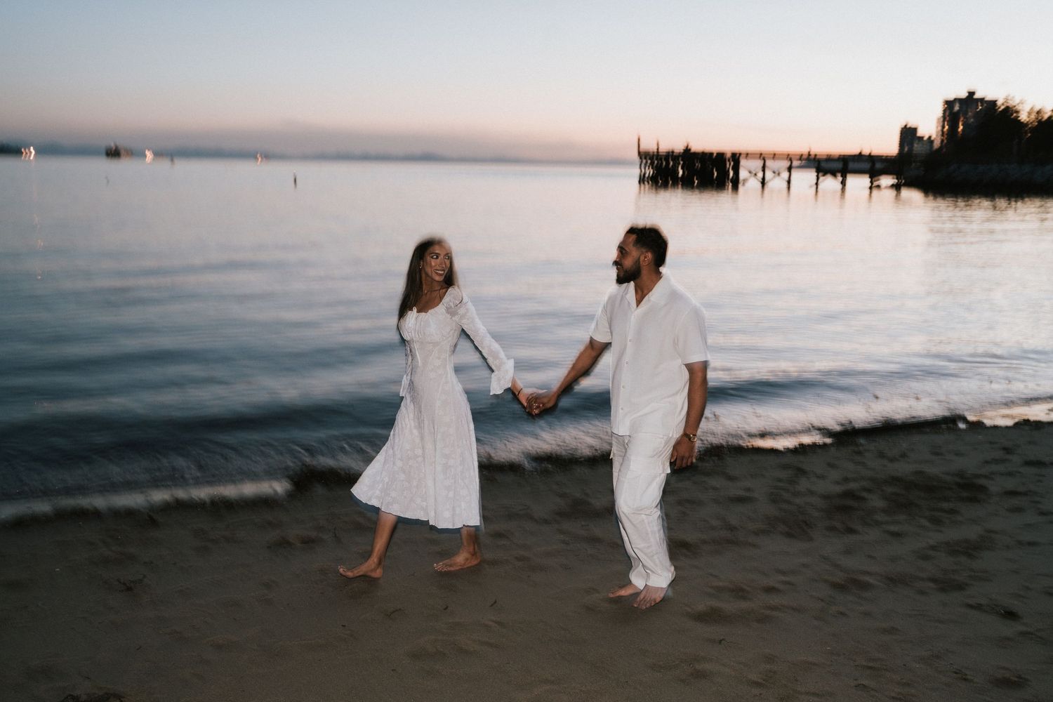 A couple in white outfits walks along a sandy beach at sunset creating a romantic silhouette against the ocean.