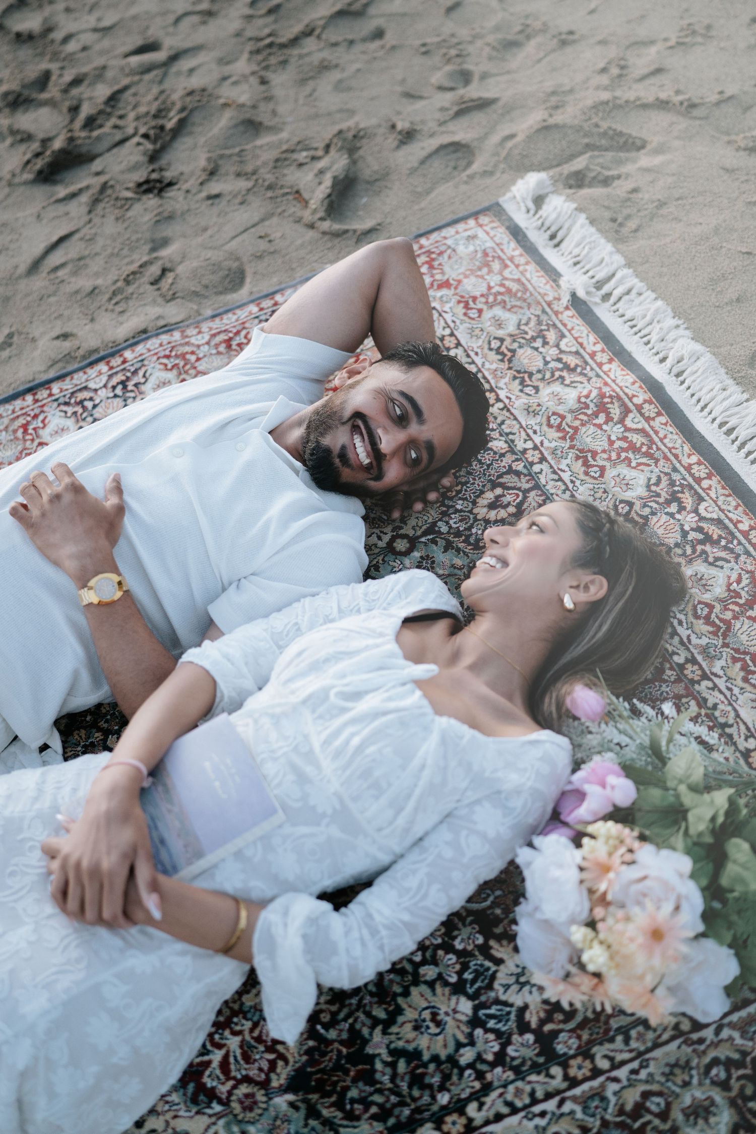 Two people in white outfits lying on a decorative rug with flowers beside them.