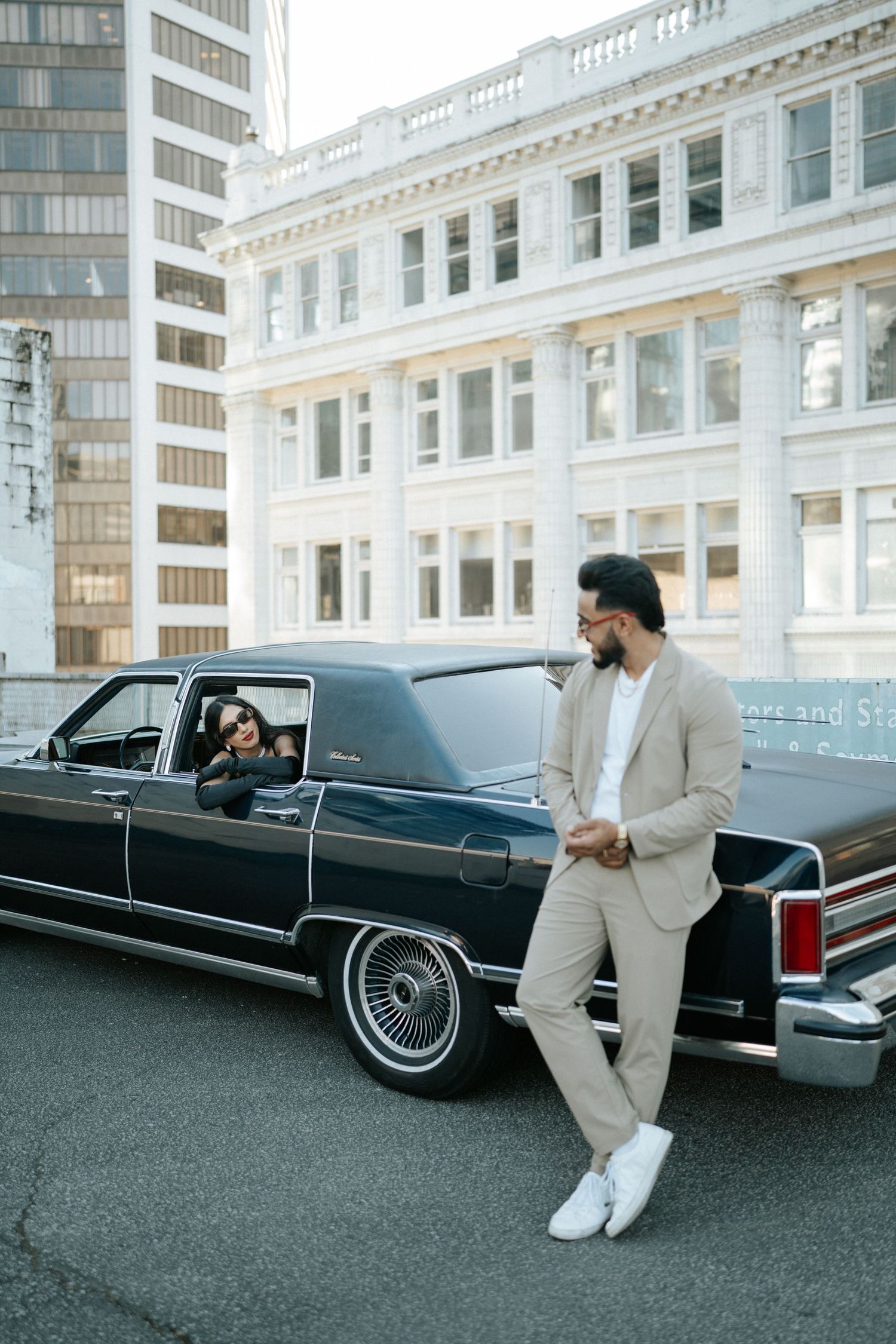 A classic black Lincoln Town Car parked in front of an urban building with someone in a light colored suit nearby.