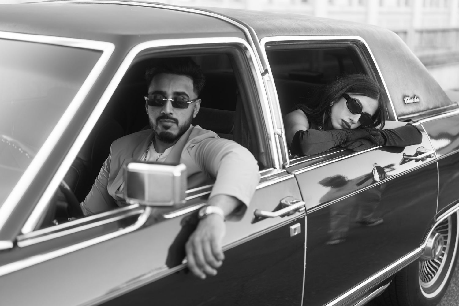Two people wearing sunglasses sit in a classic car in black and white photograph.