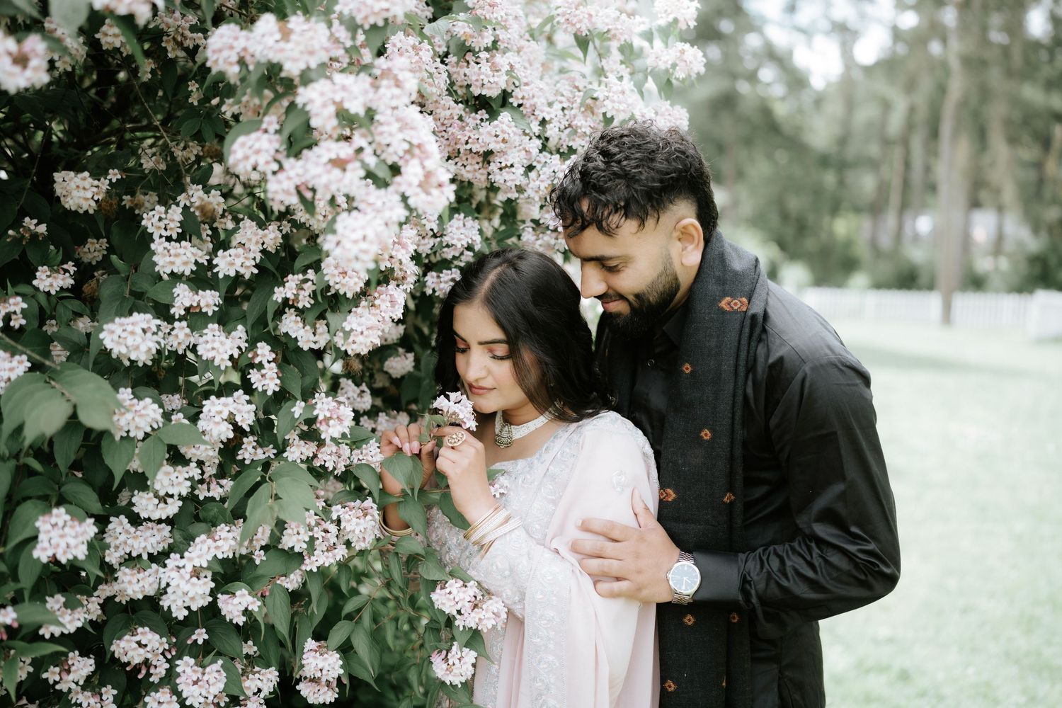 A romantic couple embraces near flowering cherry blossoms in a garden setting.