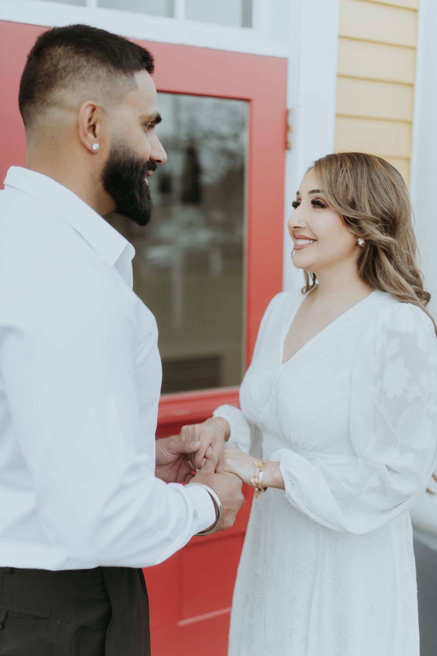 A romantic couple in white outfits share an intimate moment against a yellow building with red doors.