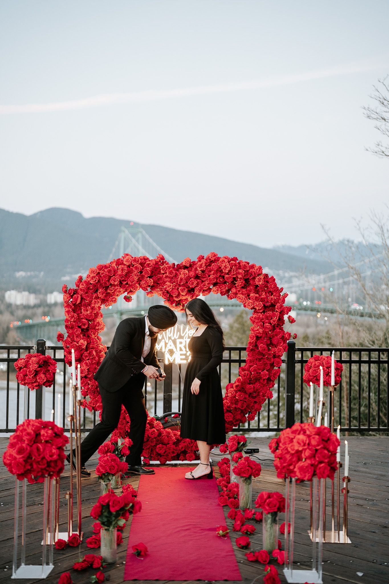 Red rose arch decorates an outdoor proposal setup with a scenic mountain and water view in the background.