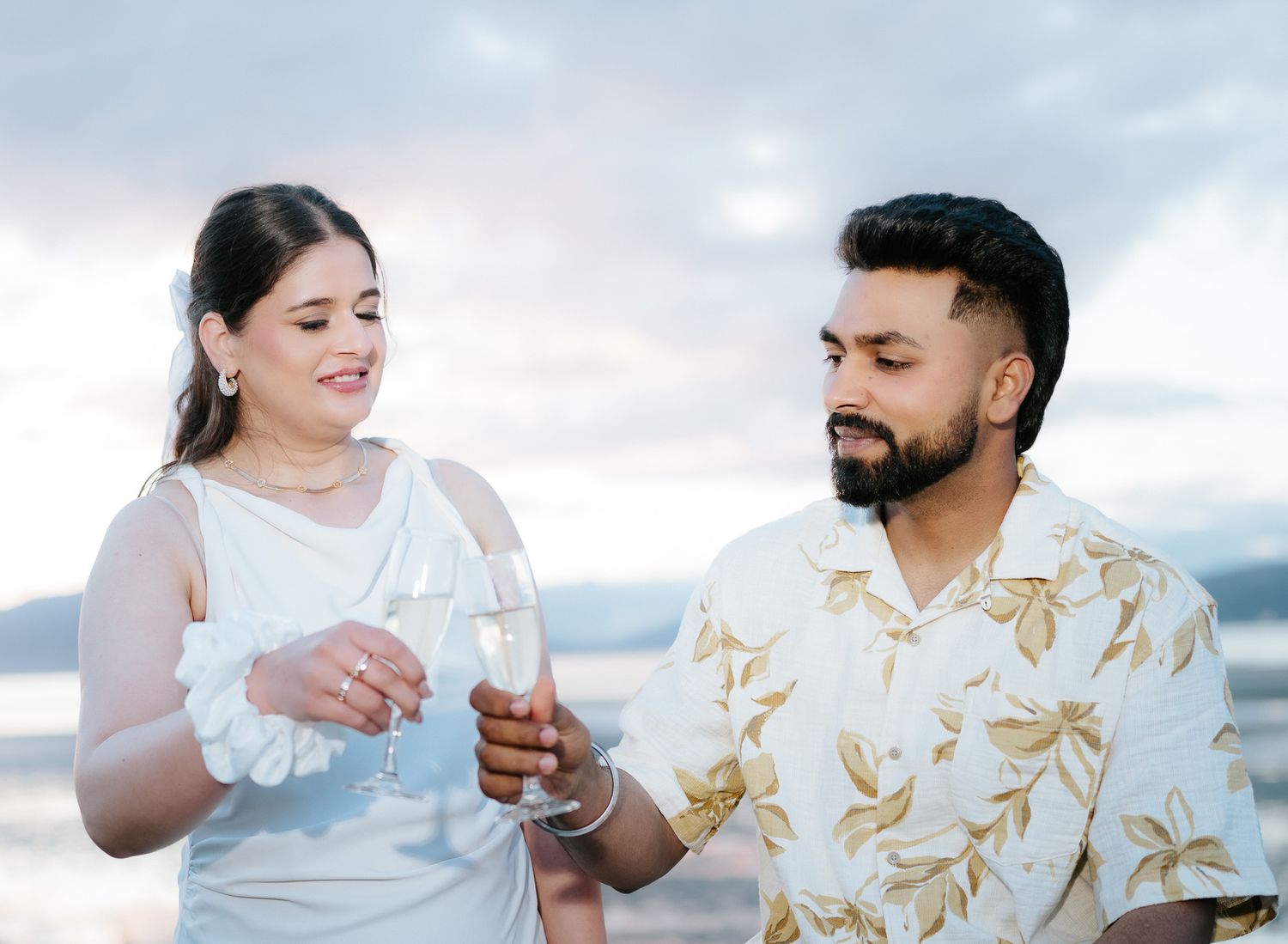 Couple toasting with champagne glasses outdoors at sunset, woman in white dress and man in floral shirt.