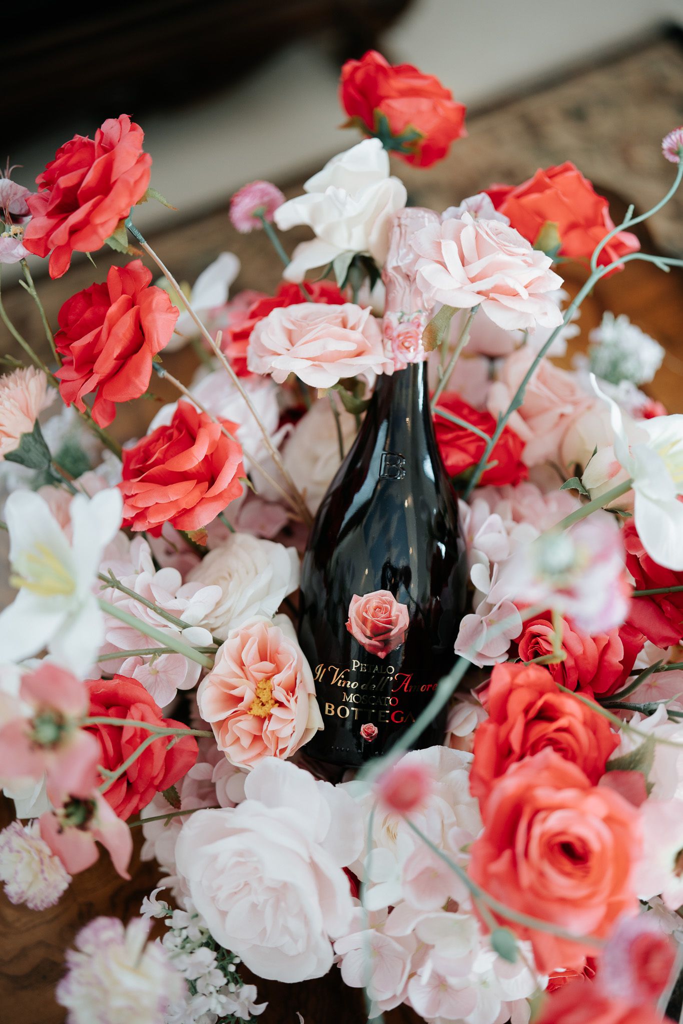 Dark wine bottle surrounded by lush pink, red, and white floral arrangement with roses and poppies.